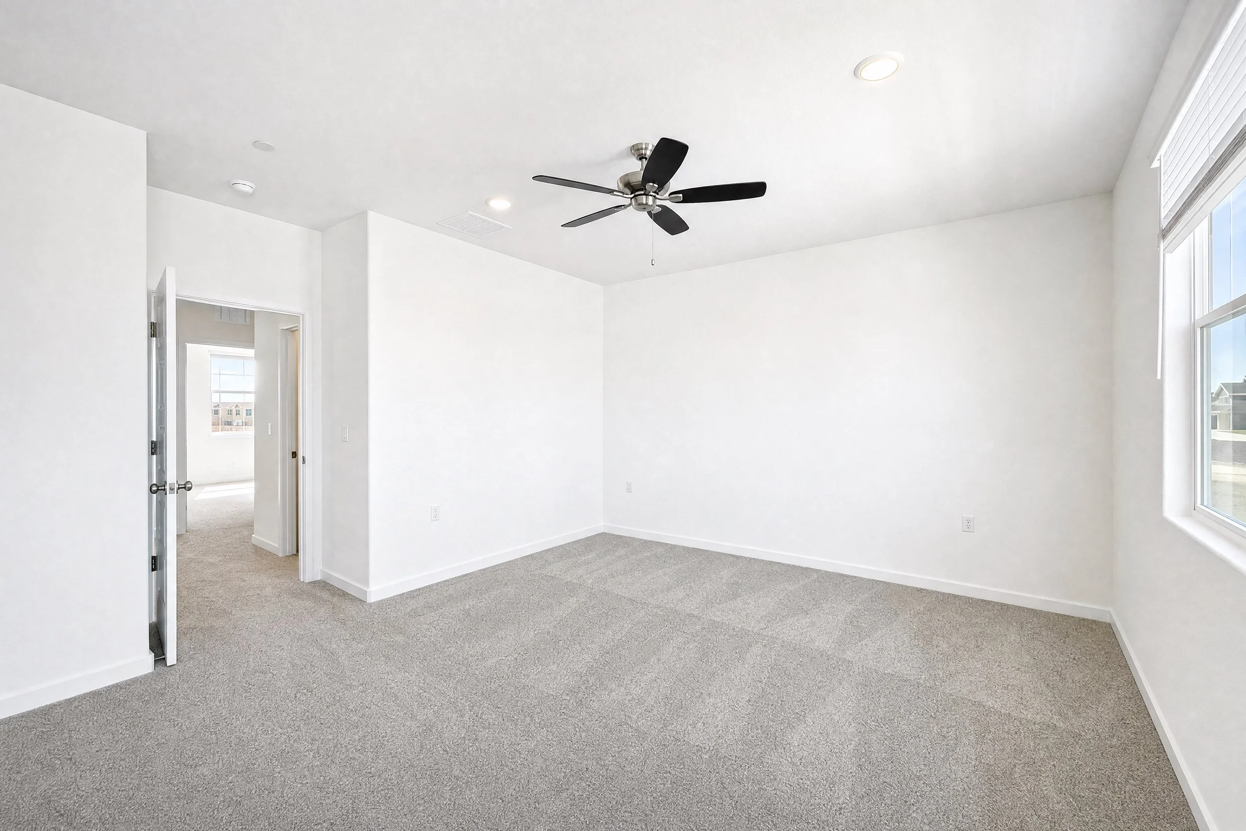 Empty white room with gray carpet, ceiling fan, large windows, and open doorway leading to another room.
