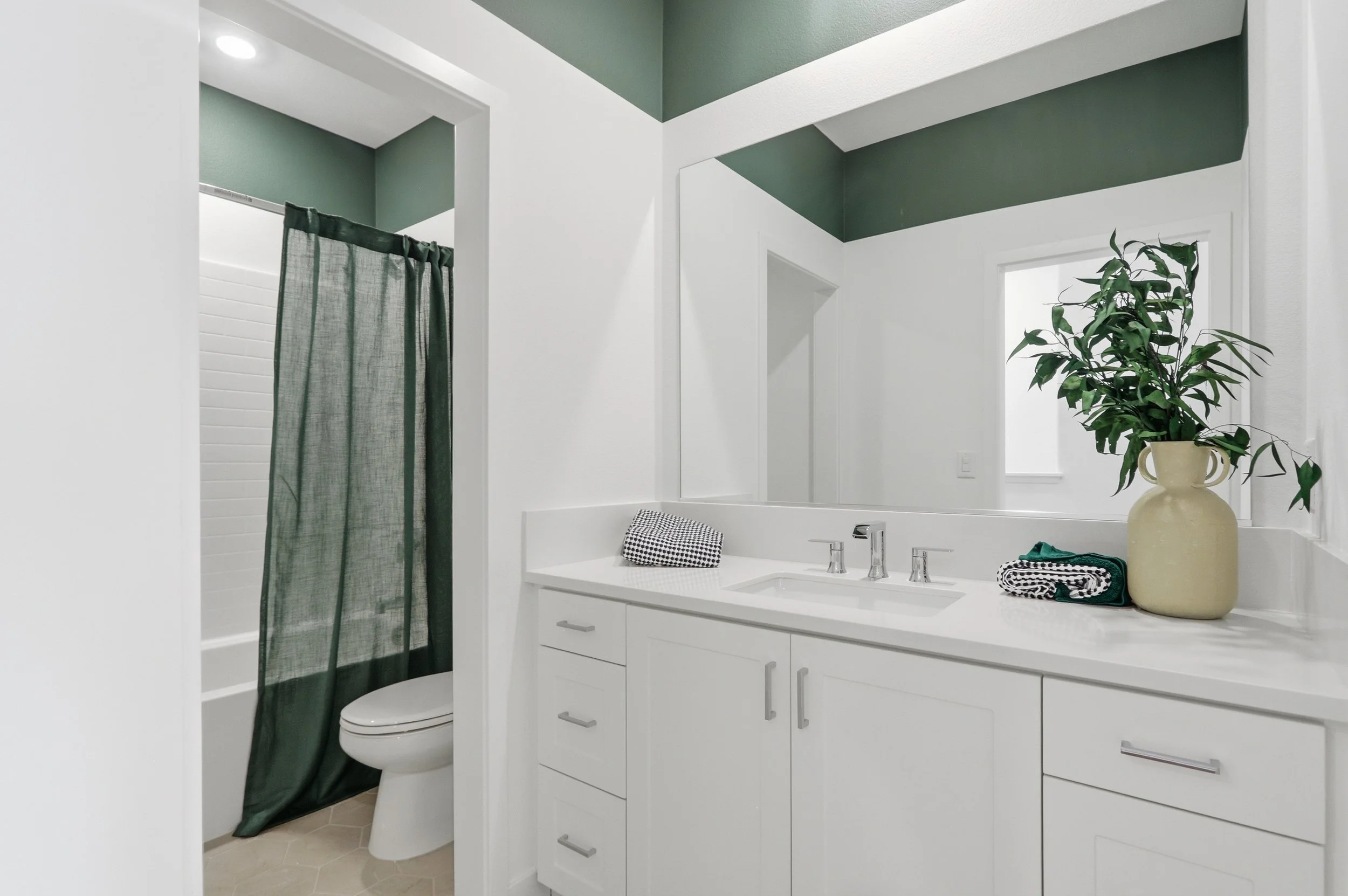 Modern bathroom with white cabinetry, large mirror, potted plant on countertop, green and white wall paint, shower with green curtain, toilet.
