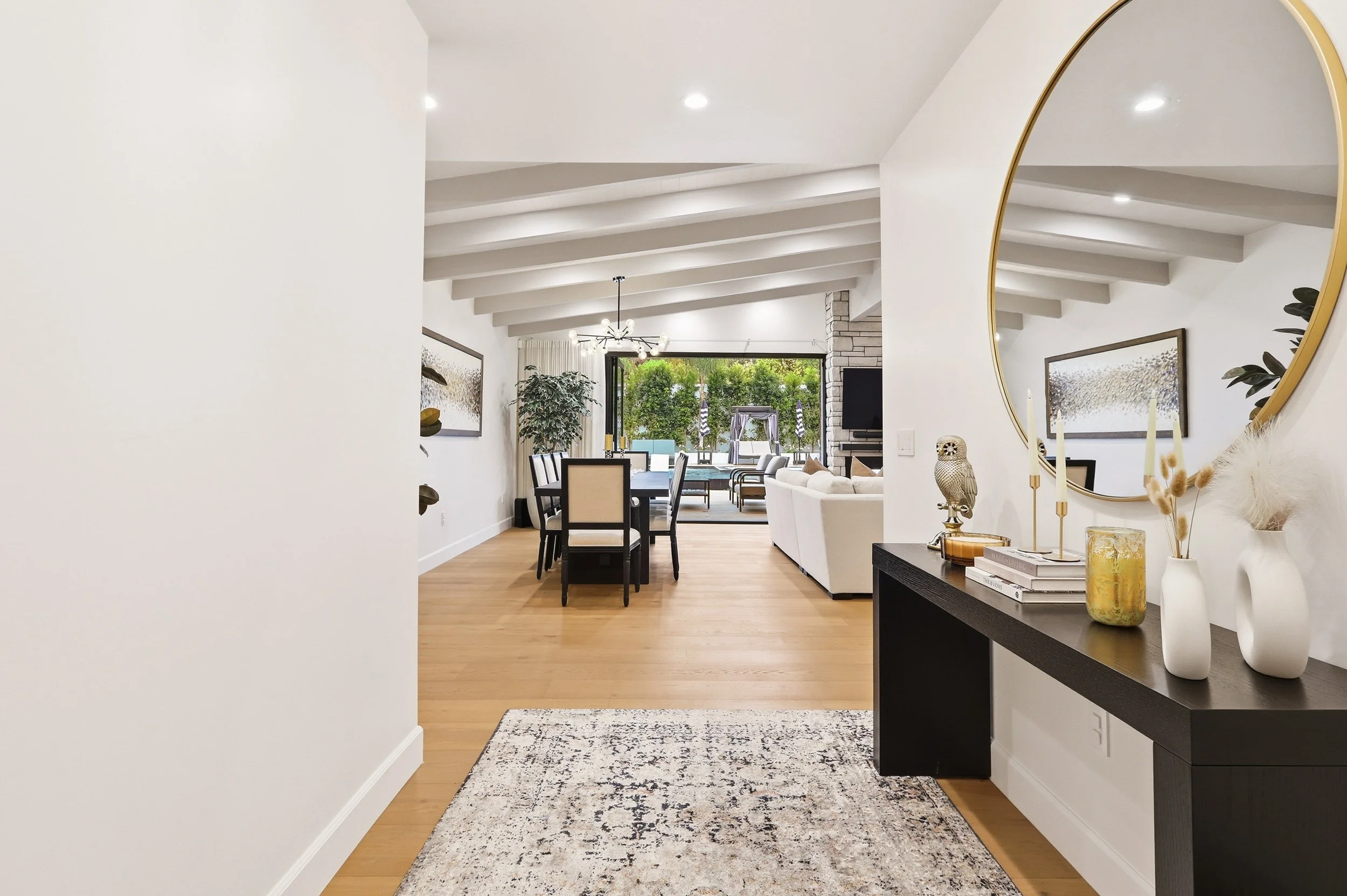 A view from a hallway into a dining and living area with modern decor, white walls, wood floors, a black side table with white vases, a large round gold-framed mirror, and a window showing outdoor greenery.