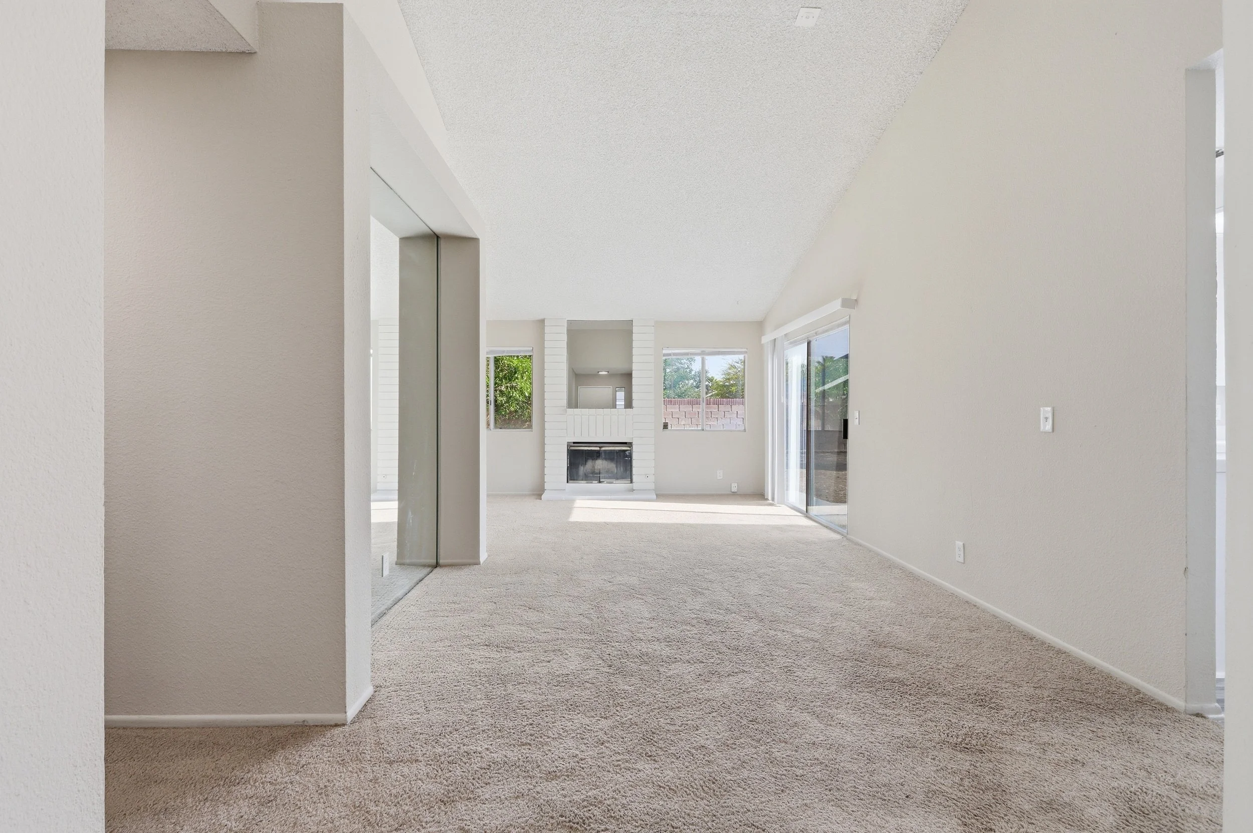 Interior of a bright, minimalist living room with white walls, a fireplace, and large windows showing an outdoor view.