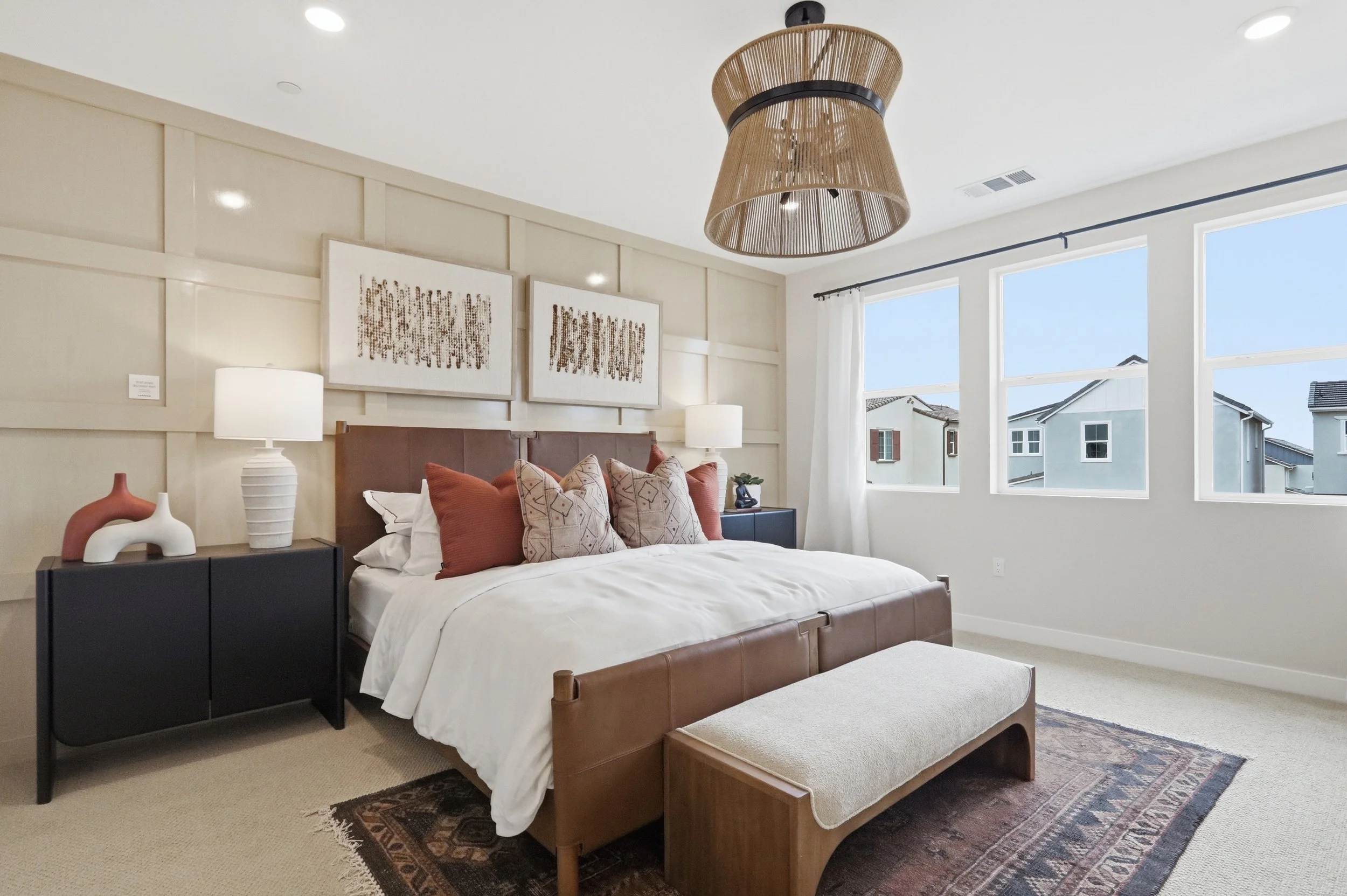 A bedroom with a bed, decorative pillows, nightstands with lamps, artwork on the wall, and large windows showing neighboring houses.