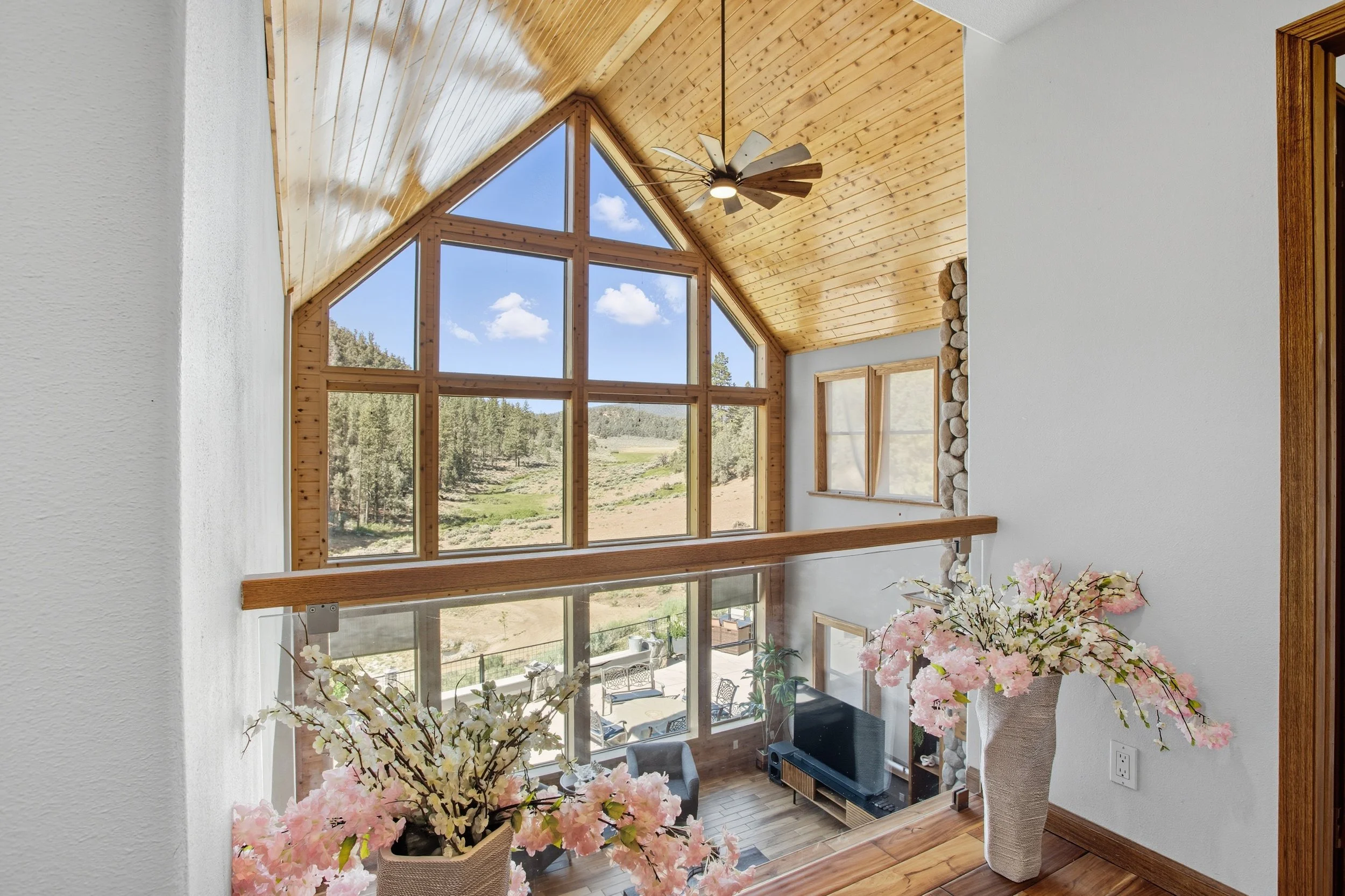 Interior view of a house with a large window wall, wooden vaulted ceiling, ceiling fan, and a view of trees and sky outside. Decor includes pink and white flowers in vases.