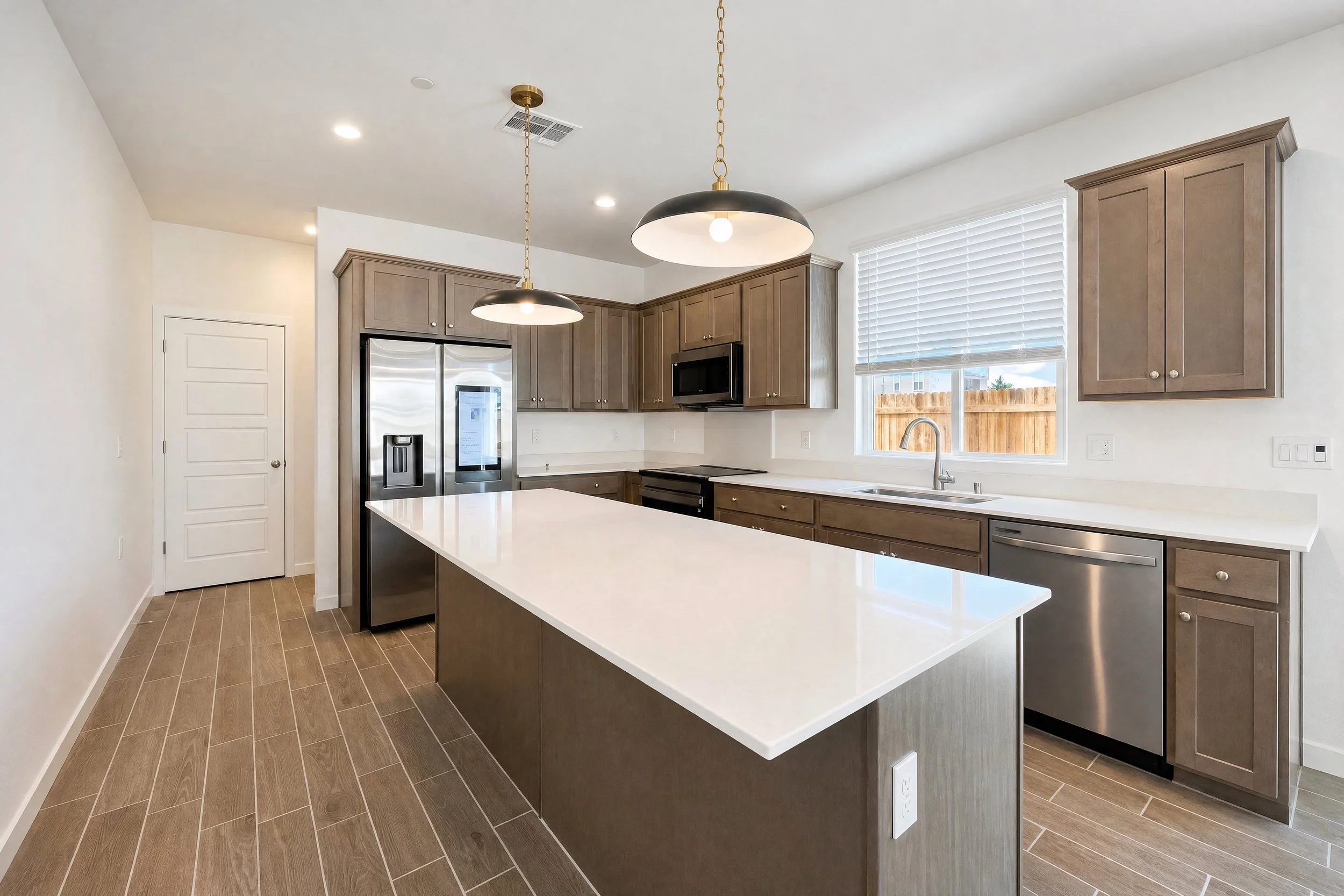 Modern kitchen with gray cabinets, white countertops, stainless steel appliances, wood-look tile floor, large window with blinds, and two black and gold pendant lights.