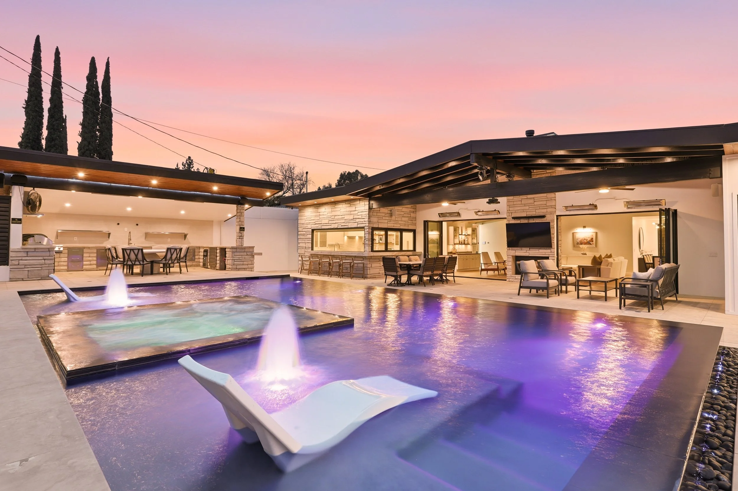 Modern outdoor pool area during sunset with lounge chairs, a hot tub, outdoor cockpit, and a seating area inside a house visible through large open doors.