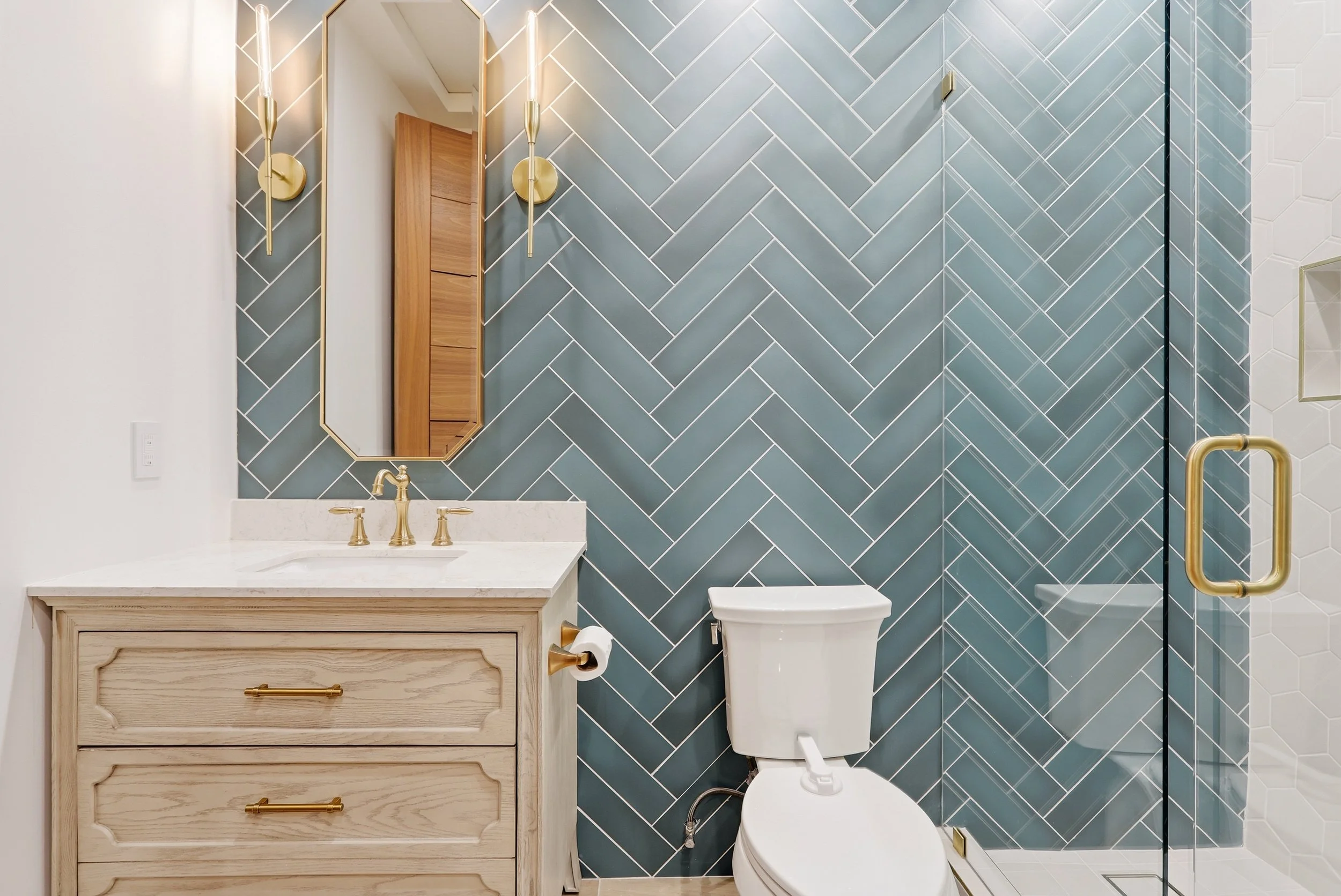 Modern bathroom with blue herringbone tiled wall, white toilet, wooden vanity with gold hardware, and a mirror with gold accents and sconces.