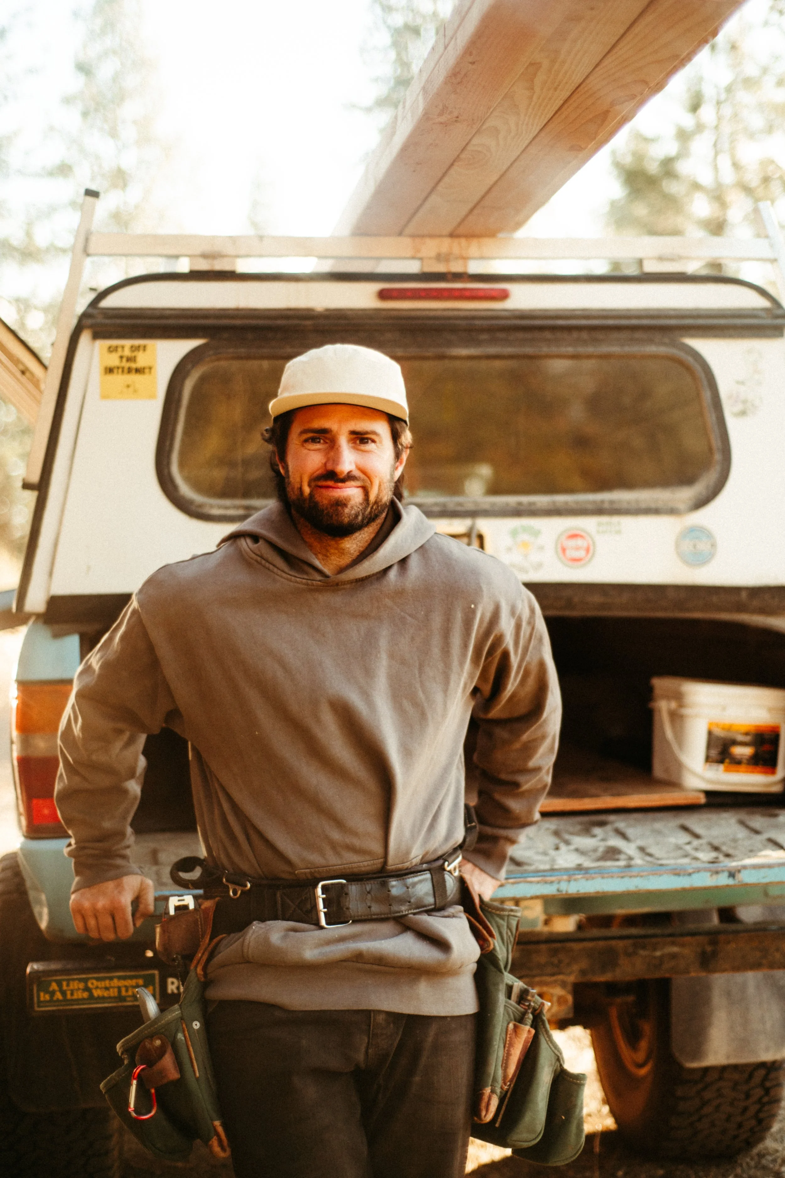 A man with a beard and mustache wearing a beige cap and gray hoodie, standing in front of a trailer with tools and equipment. There is a yellow sign that says "Get off the internet" and a sticker of a red crossed-out camera. The scene is outdoors with trees and sunlight in the background.