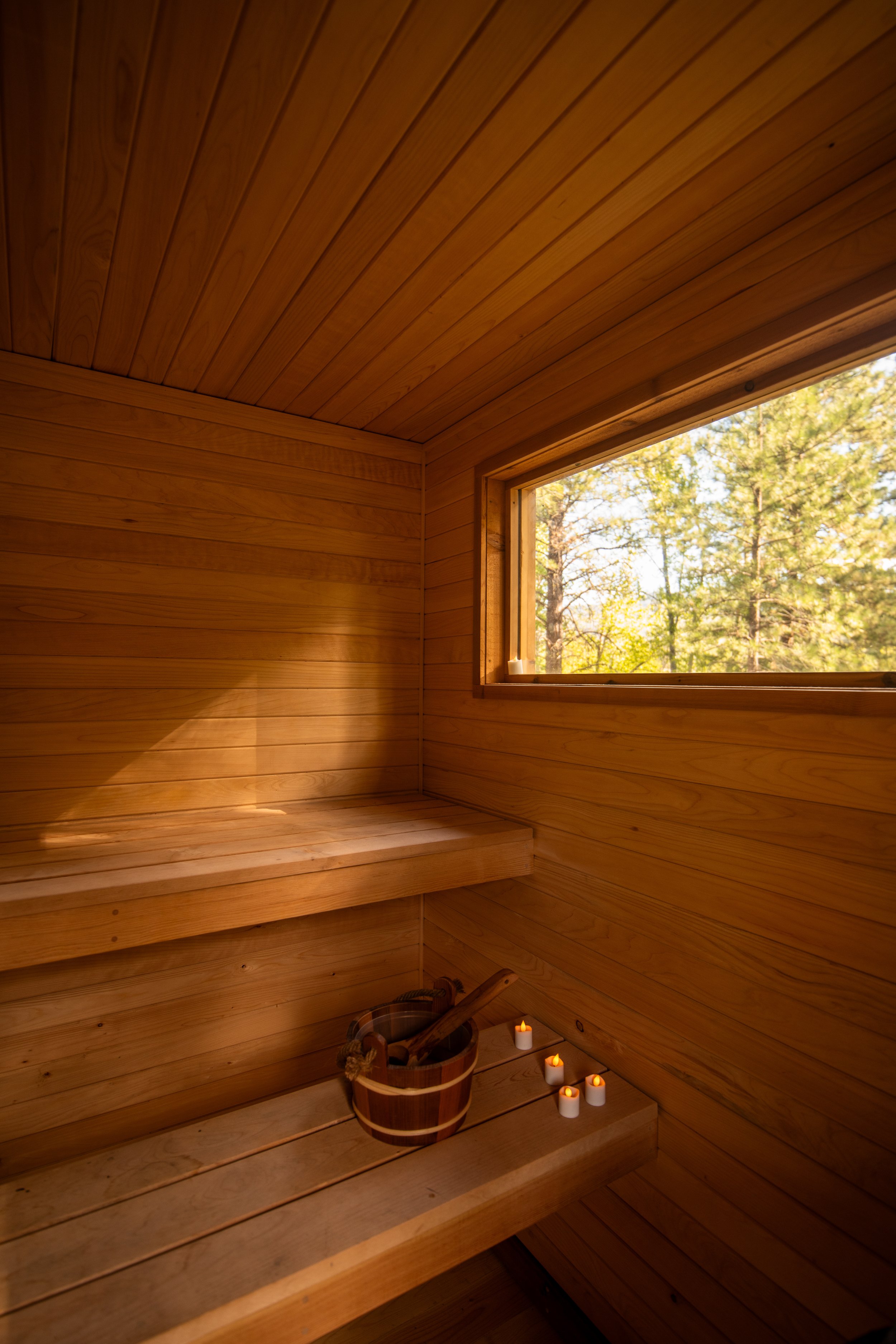 Wood-paneled sauna interior with a window showing trees outside, a bucket with a wooden ladle, and candles on the floor.