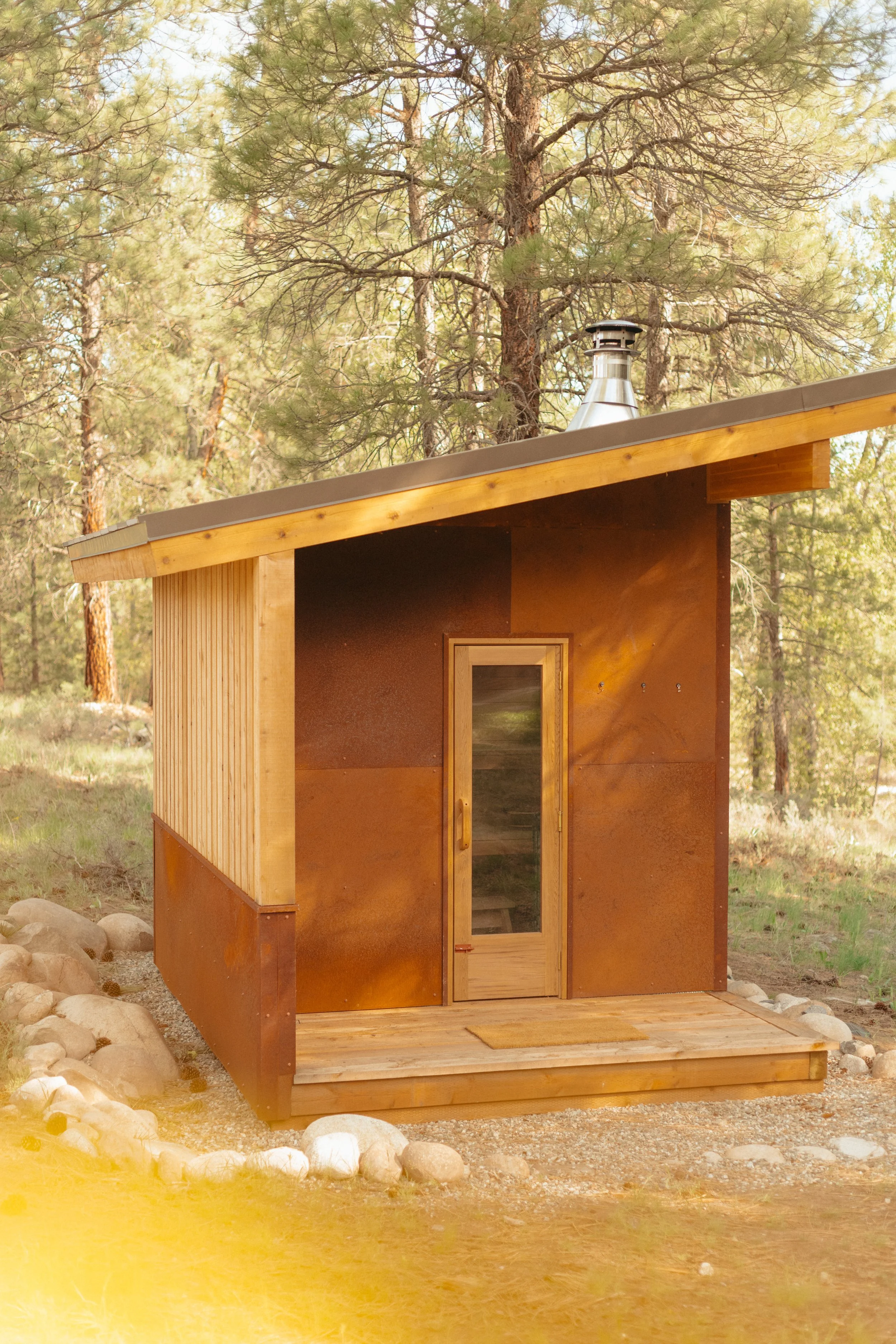Custom sundog sauna with a pitched roof, large window, and a wooden deck in a forested area with pine trees in the background.