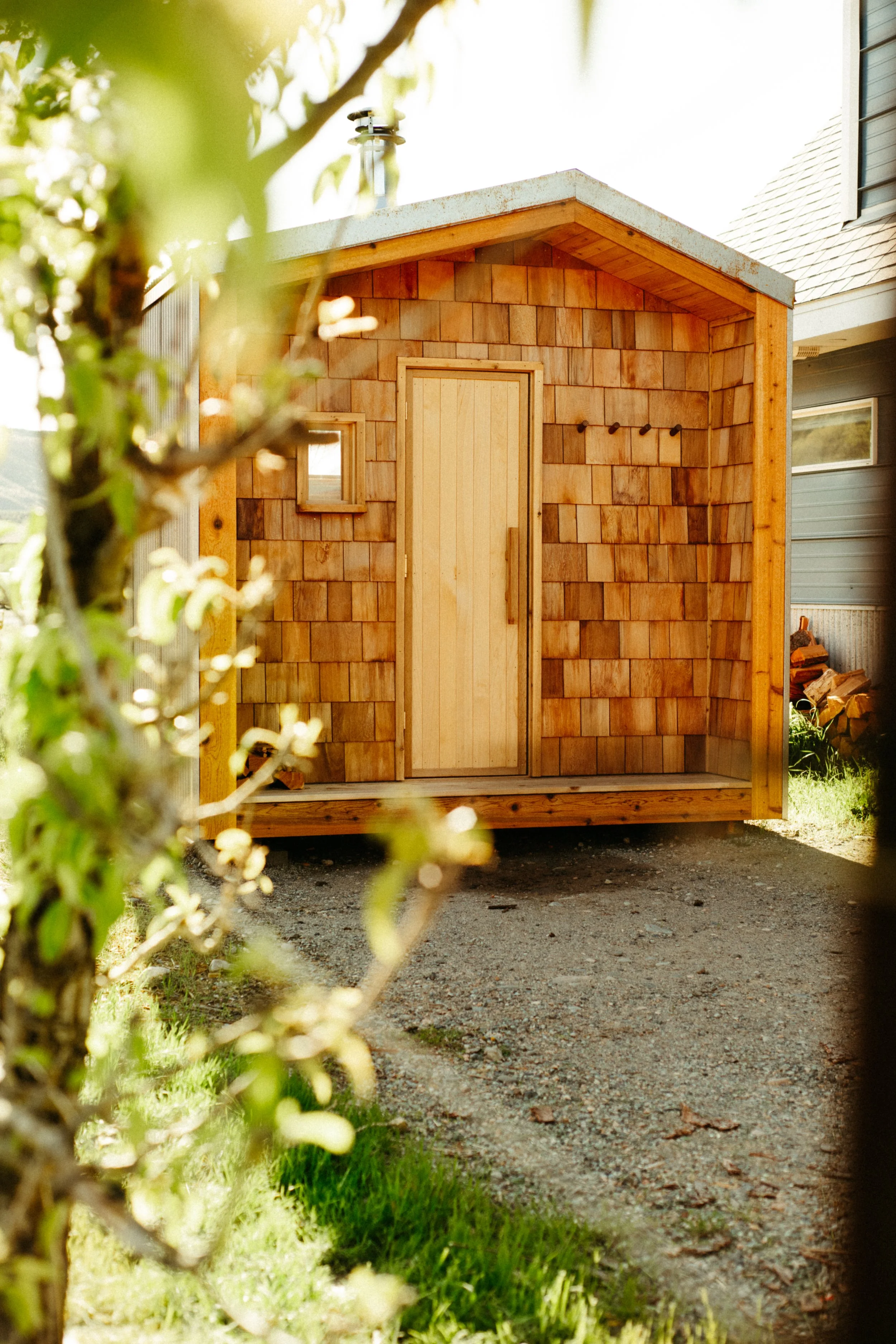 A small custom wooden sauna with a gabled roof and a closed door, surrounded by green grass in the pacific northwest, with a dirt path leading to it.