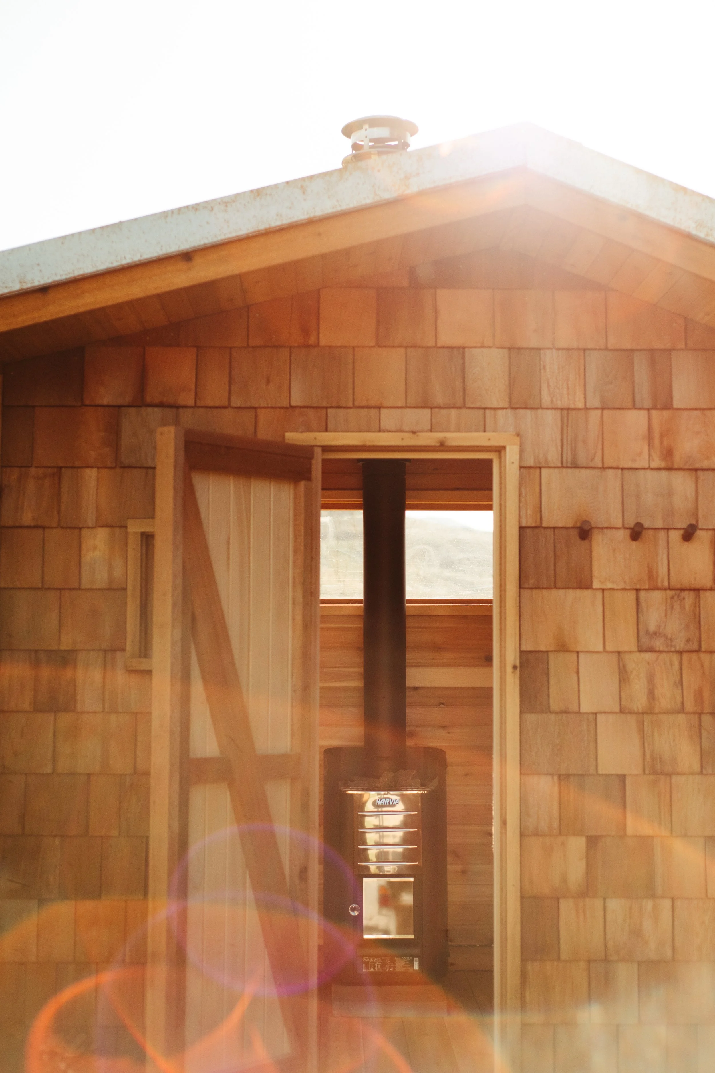 Interior view of a custom sauna with an open door, a wood stove and a window with a view of a landscape outside.