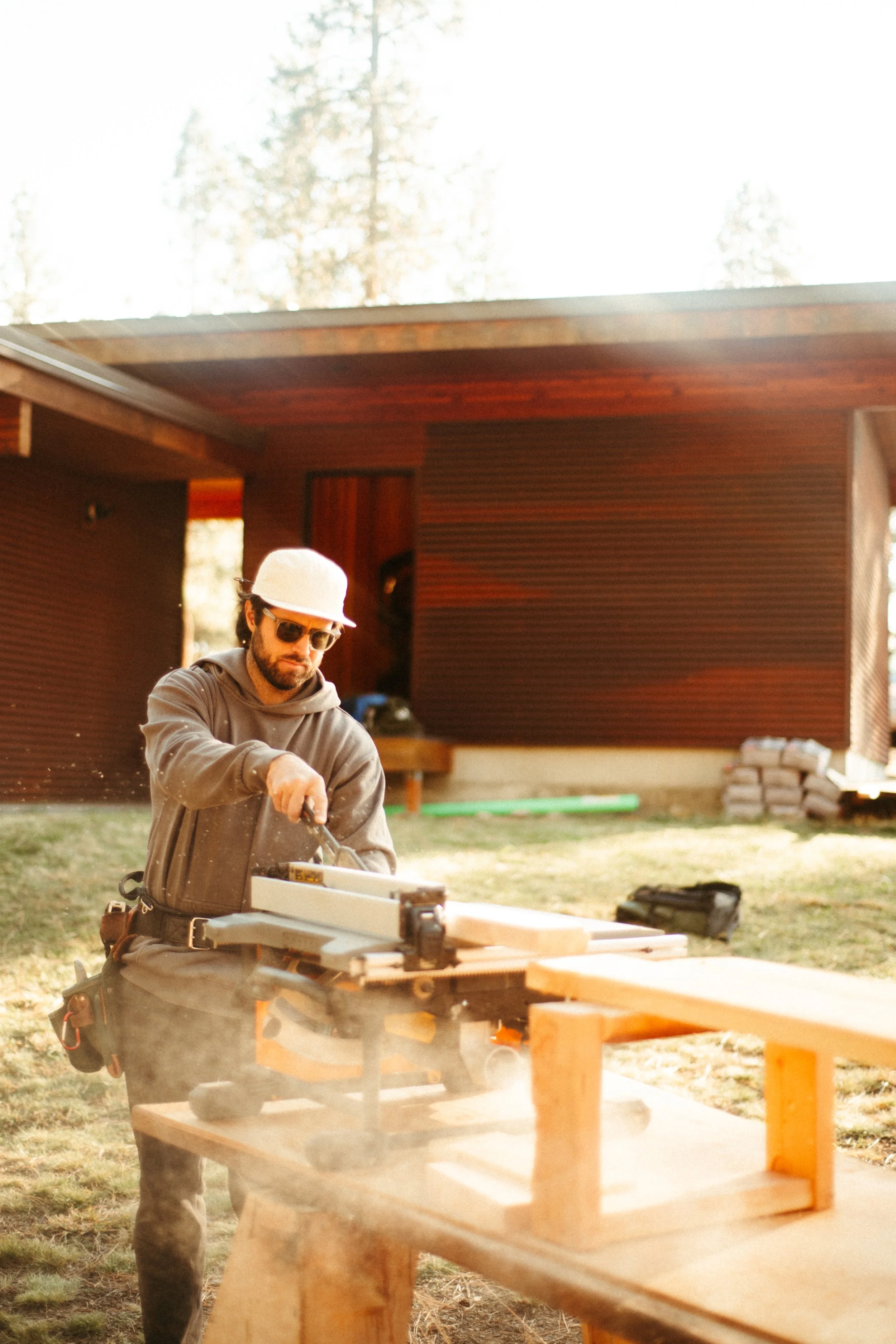 Man wearing sunglasses, a white hat, and work clothes is woodworking outdoors at a construction site with a partially built custom sauna in the foreground.
