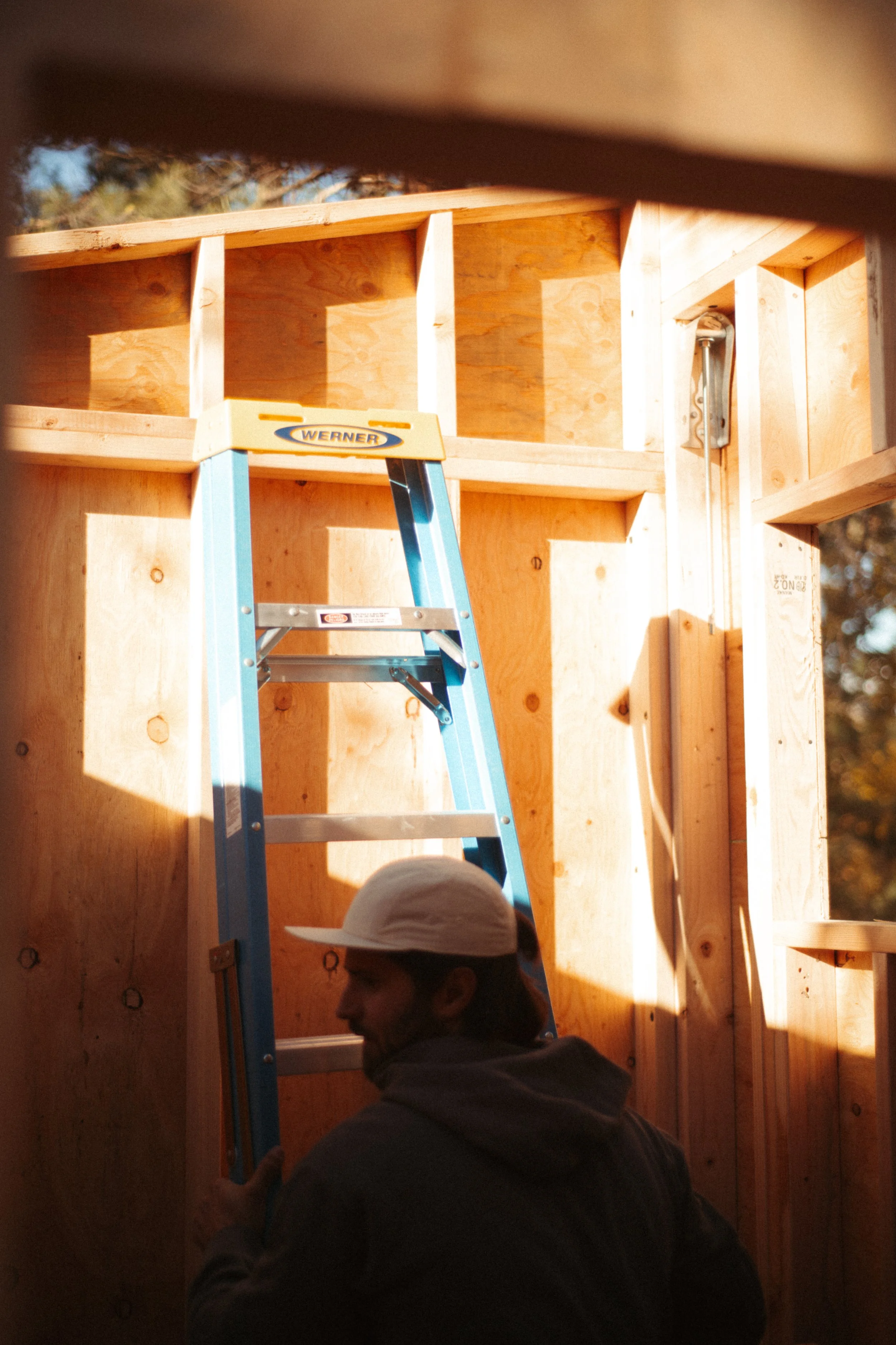 Person wearing a white hat working on a wooden structure, with a blue ladder inside a partially constructed custom sauna building.