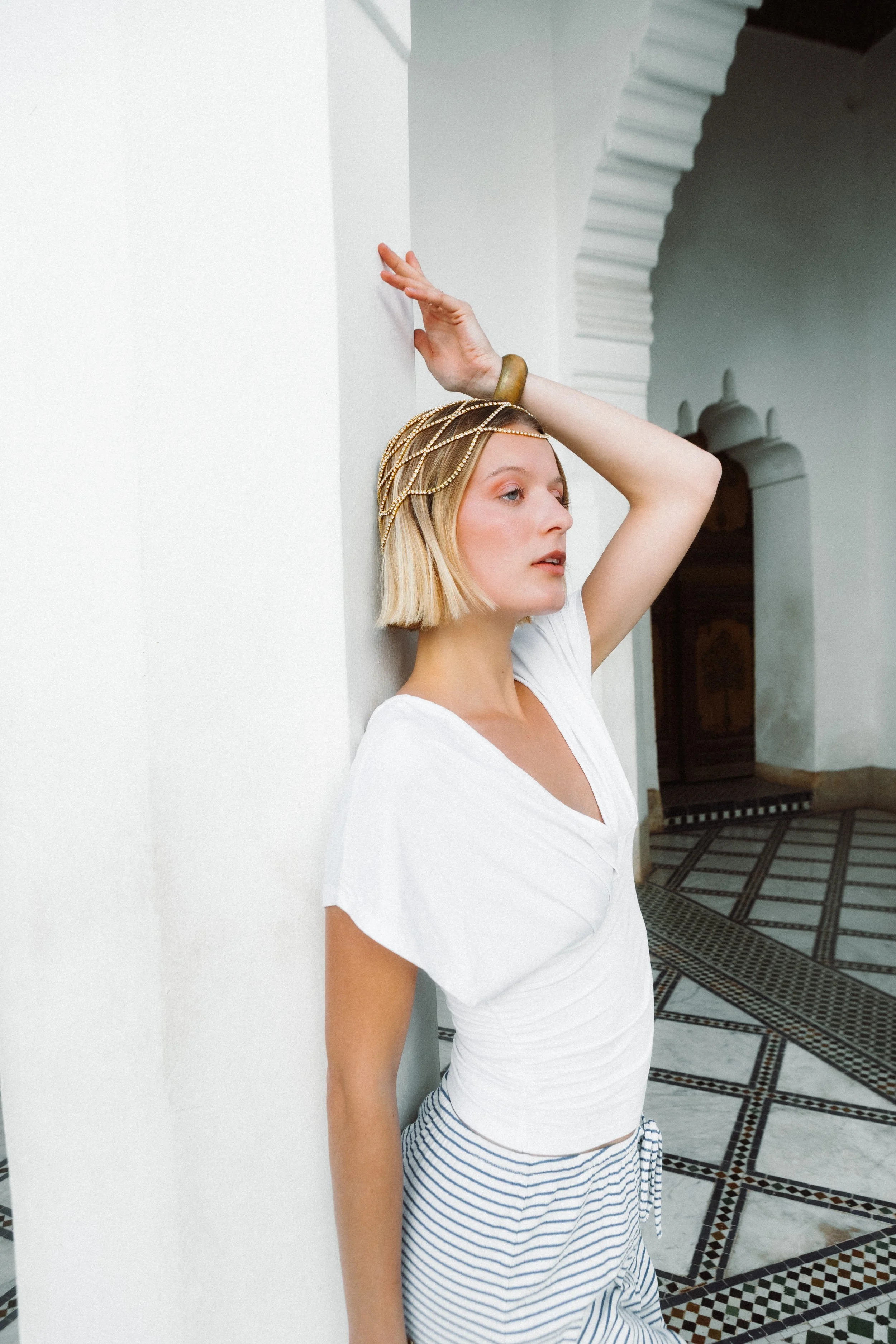 A young woman with blonde hair, wearing a white t-shirt, striped skirt, gold headpiece, and a bracelet, leaning against a white wall in a room with decorative flooring and an archway.