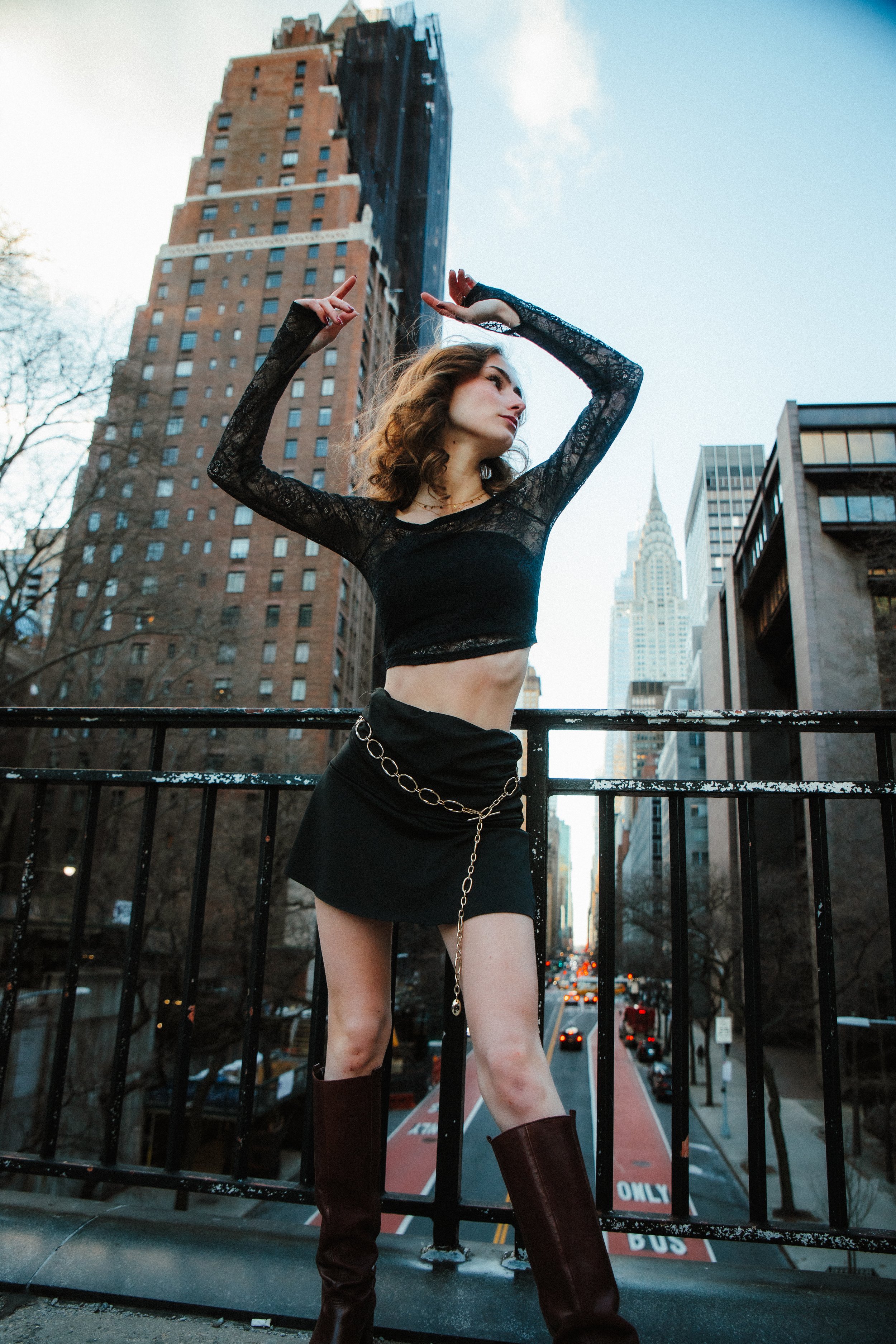A woman standing on a city bridge in front of tall buildings, wearing a black lace crop top, black skirt, and knee-high boots, posed with his arms raised above her head and looking to the side.