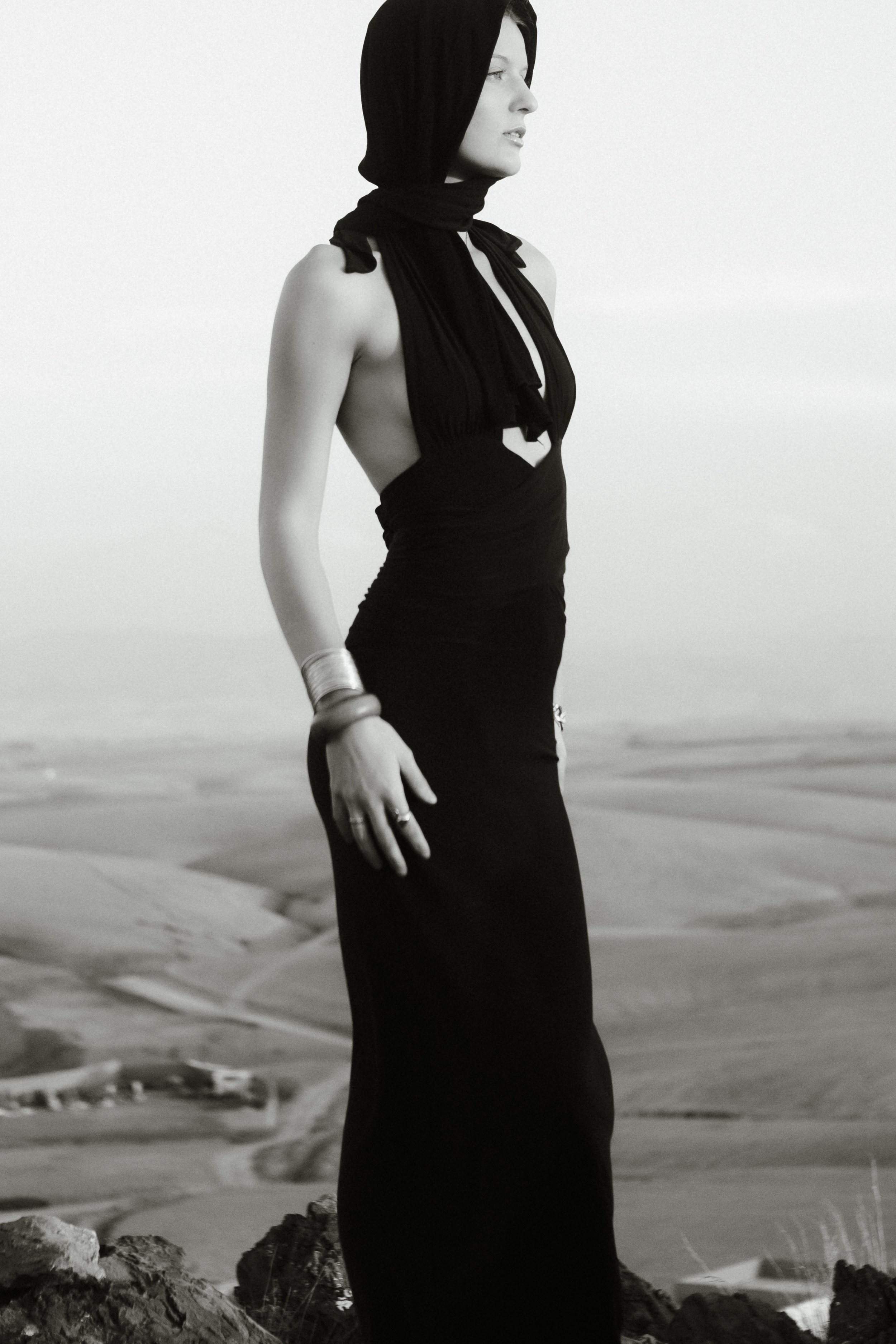 A woman stands on the beach facing right, wearing a long black dress with an open side and a black hood covering her head.