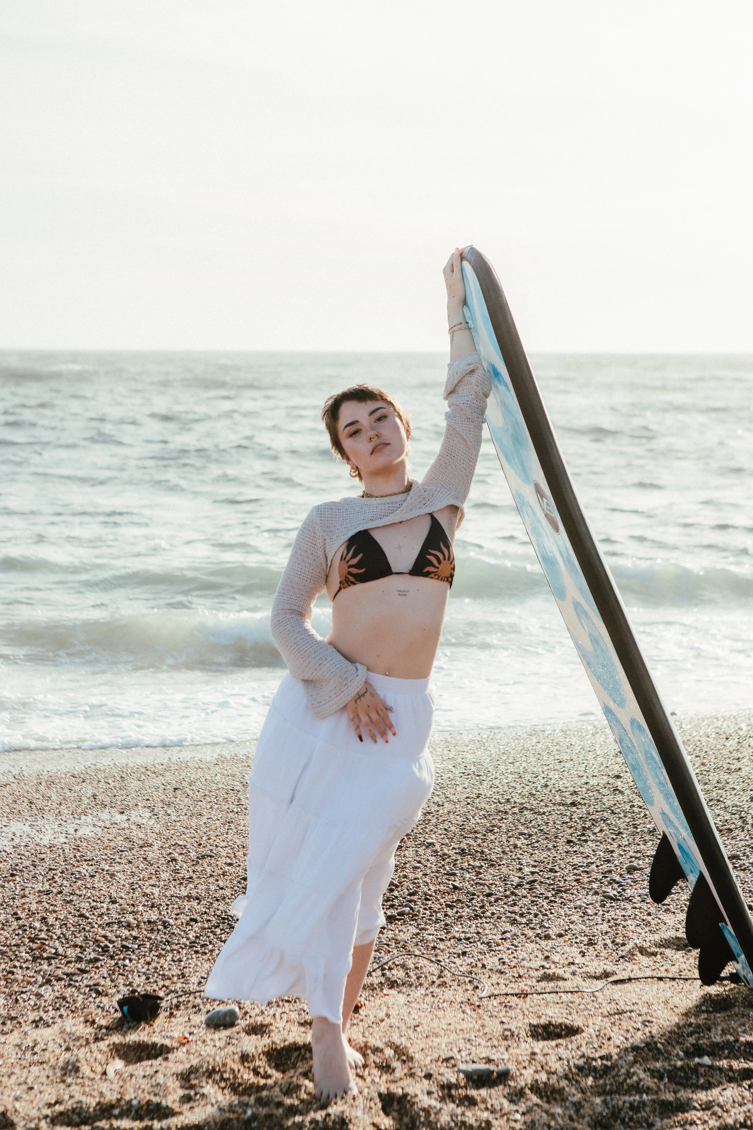 A woman standing on a beach holding a surfboard, with ocean waves in the background and the sky. She is wearing a black bikini top with a orange design, a light sweater, and white pants.