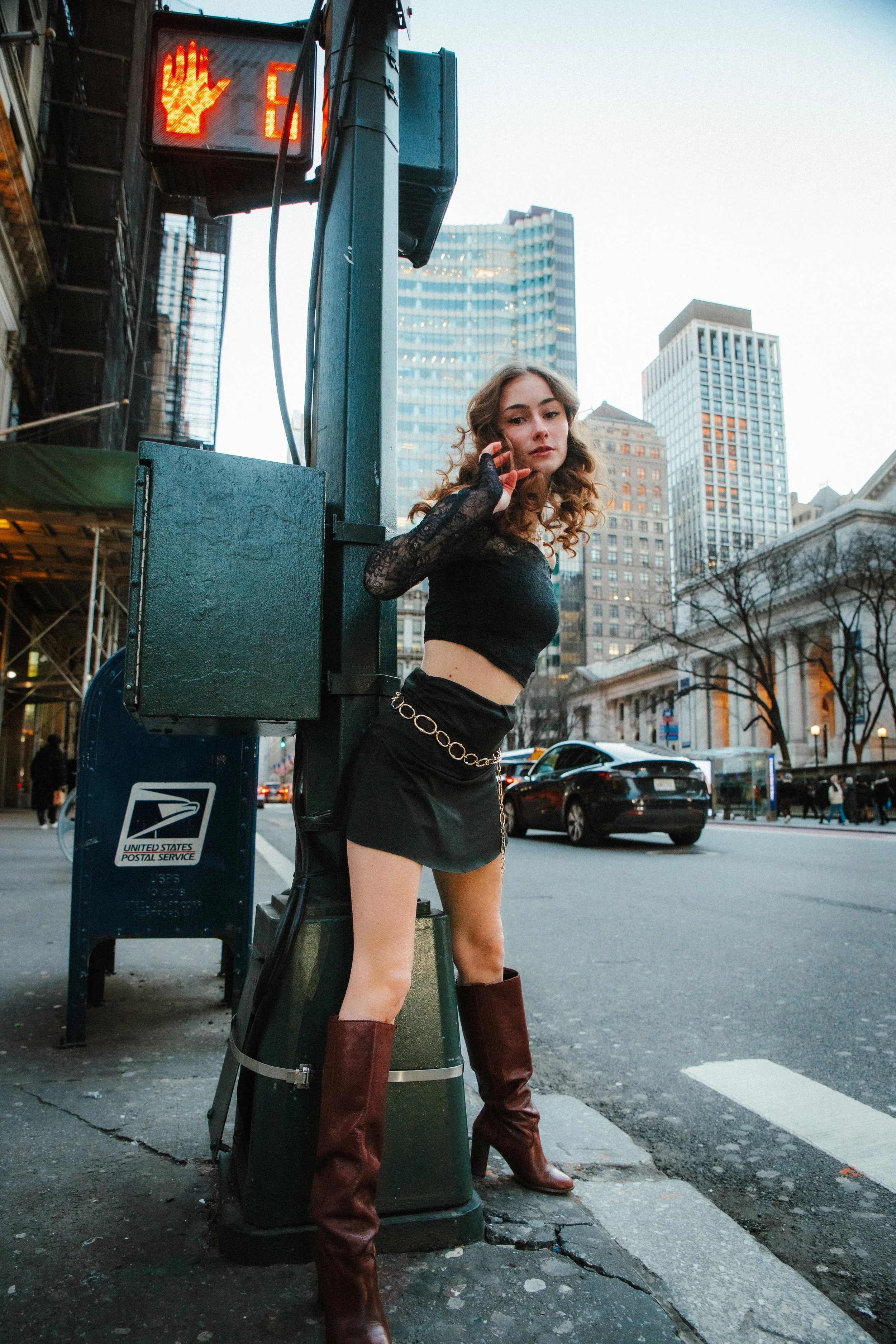A young woman with curly hair wearing a black crop top, black skirt with a chain belt, and tall brown boots, stands on the street near a mailbox and traffic light with a red hand indicator, in an urban cityscape with tall buildings in the background 