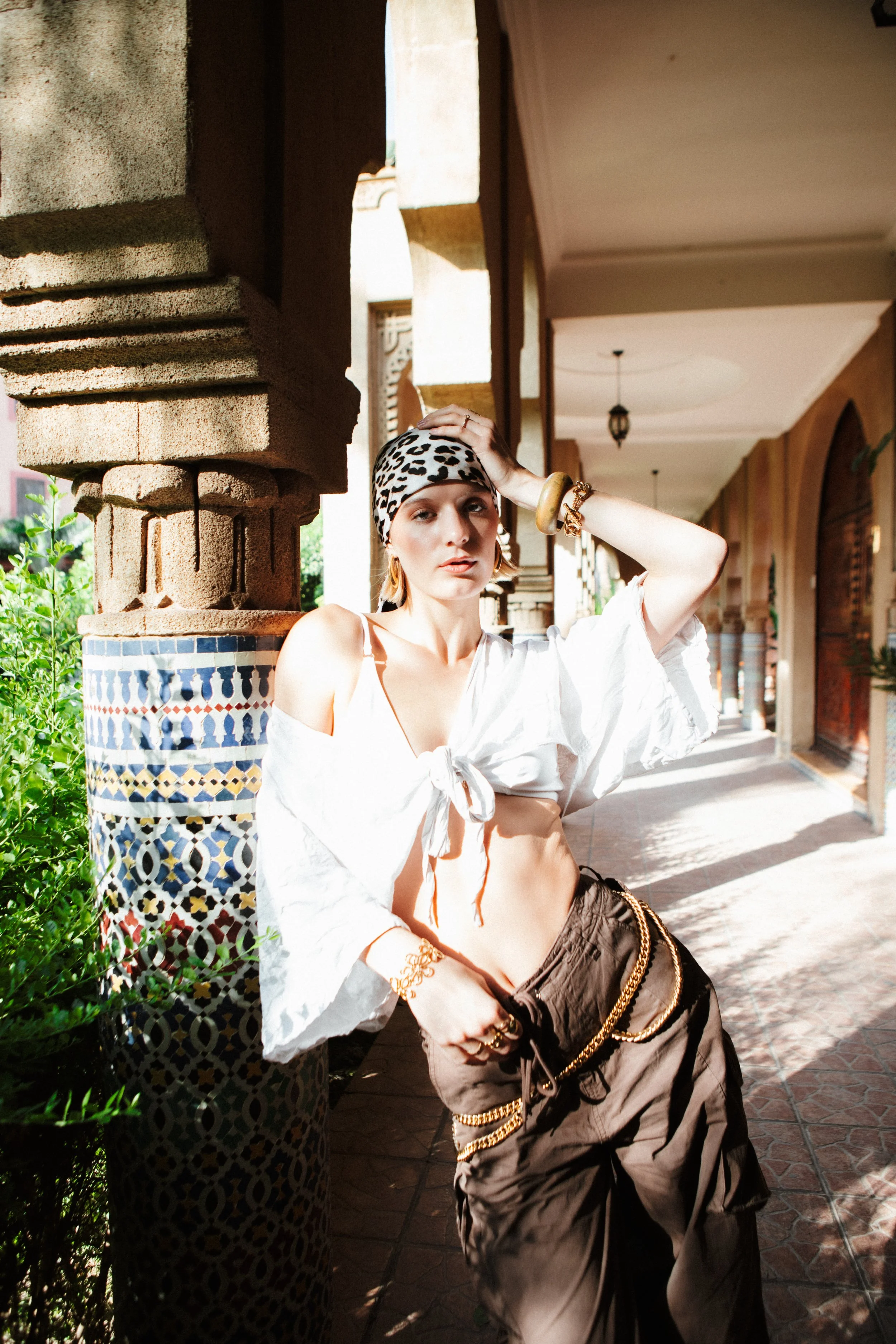 A woman leaning against a decorative column outdoors, wearing a leopard print headscarf, white off-shoulder blouse, brown pants with gold chains, and accessories, standing in a shaded corridor with arches and hanging lanterns.