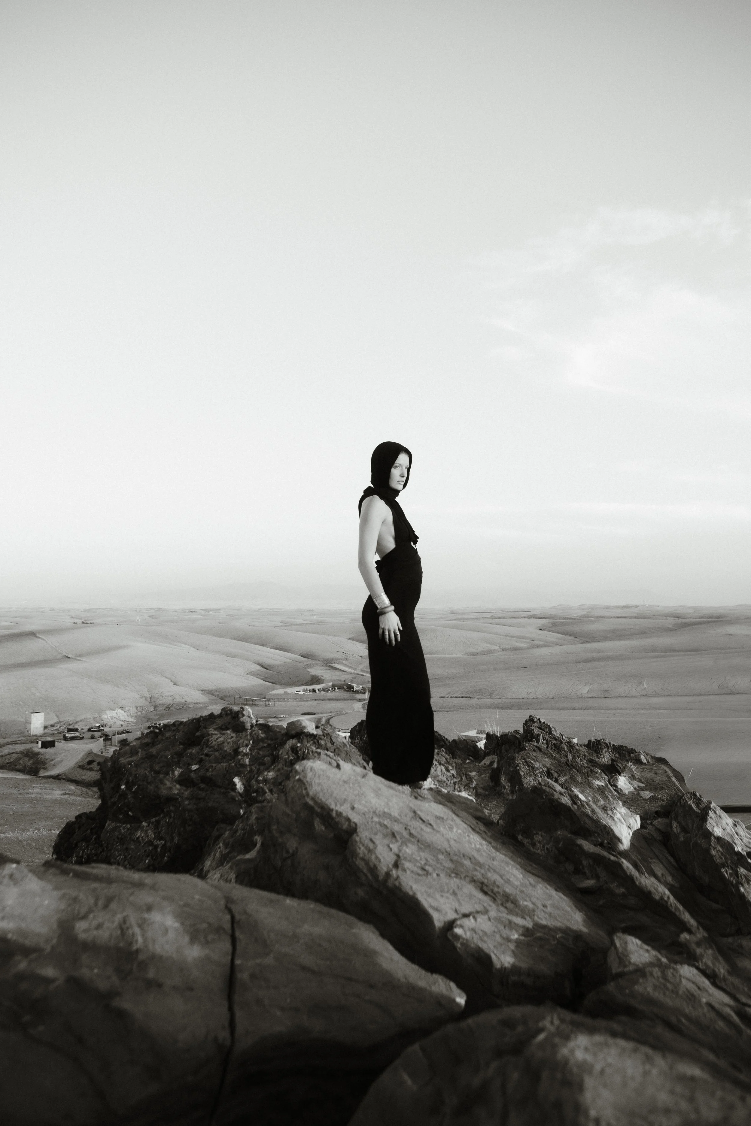 A black and white photo of a woman in a long dress and head covering, standing on rocky terrain, overlooking an open landscape with no visible buildings or trees and a cloudy sky.