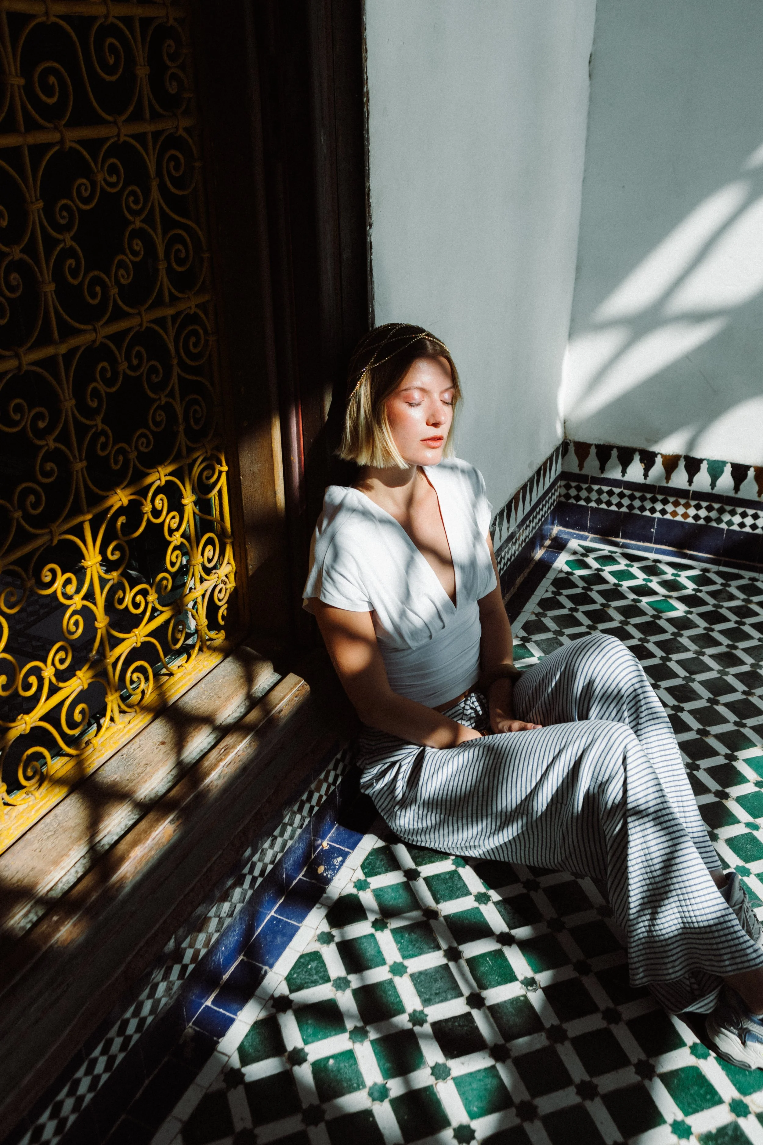 A woman with short blond hair wearing a white top and striped pants is sitting on the floor with her eyes closed in a room with Moroccan-style patterned tiles on the floor and wall, near a wooden door with an ornate yellow metal grille, and a white w