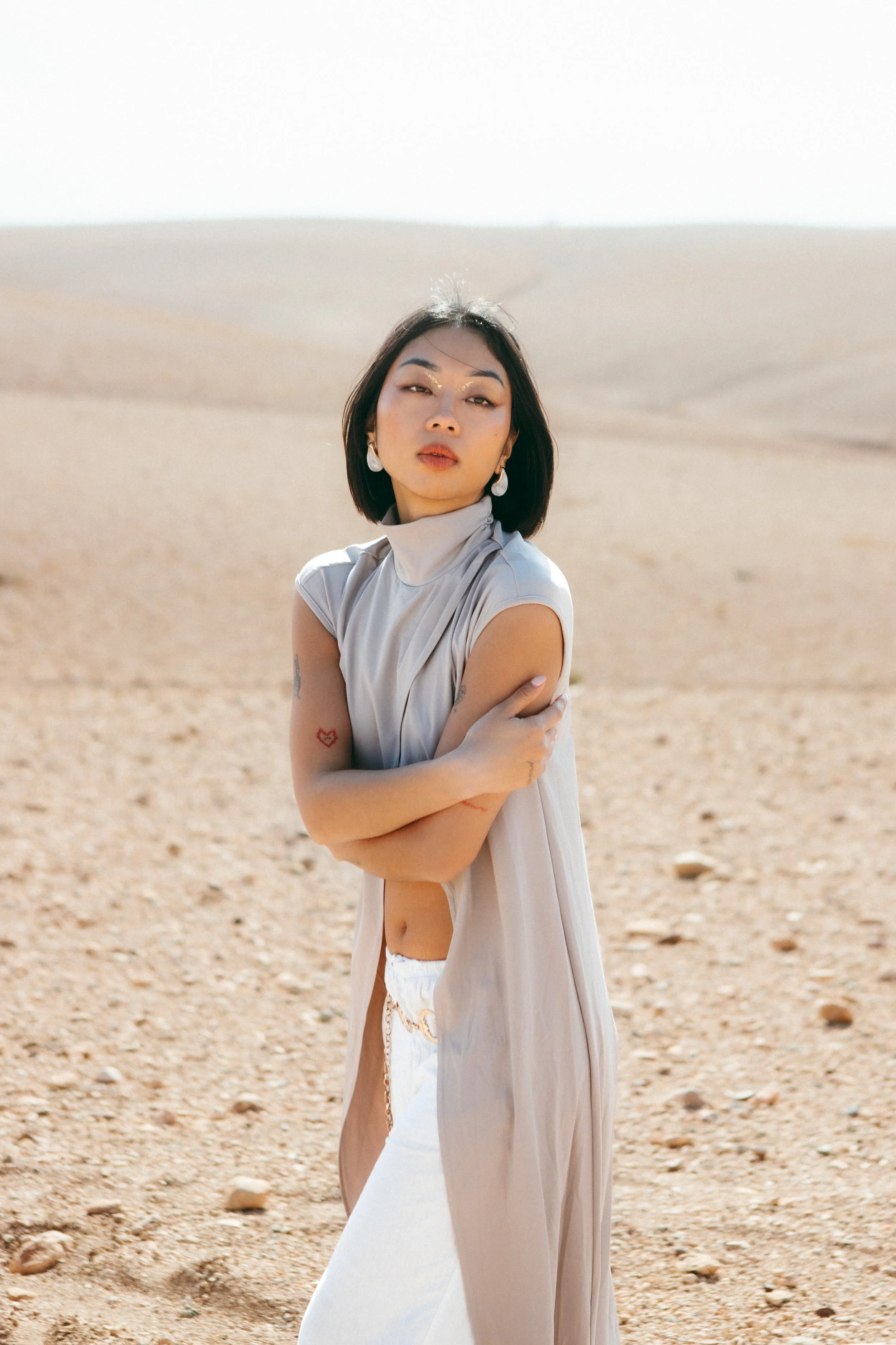 A woman with short dark hair standing in a desert, wearing a light-colored sleeveless top, with her arms crossed and looking at the camera.