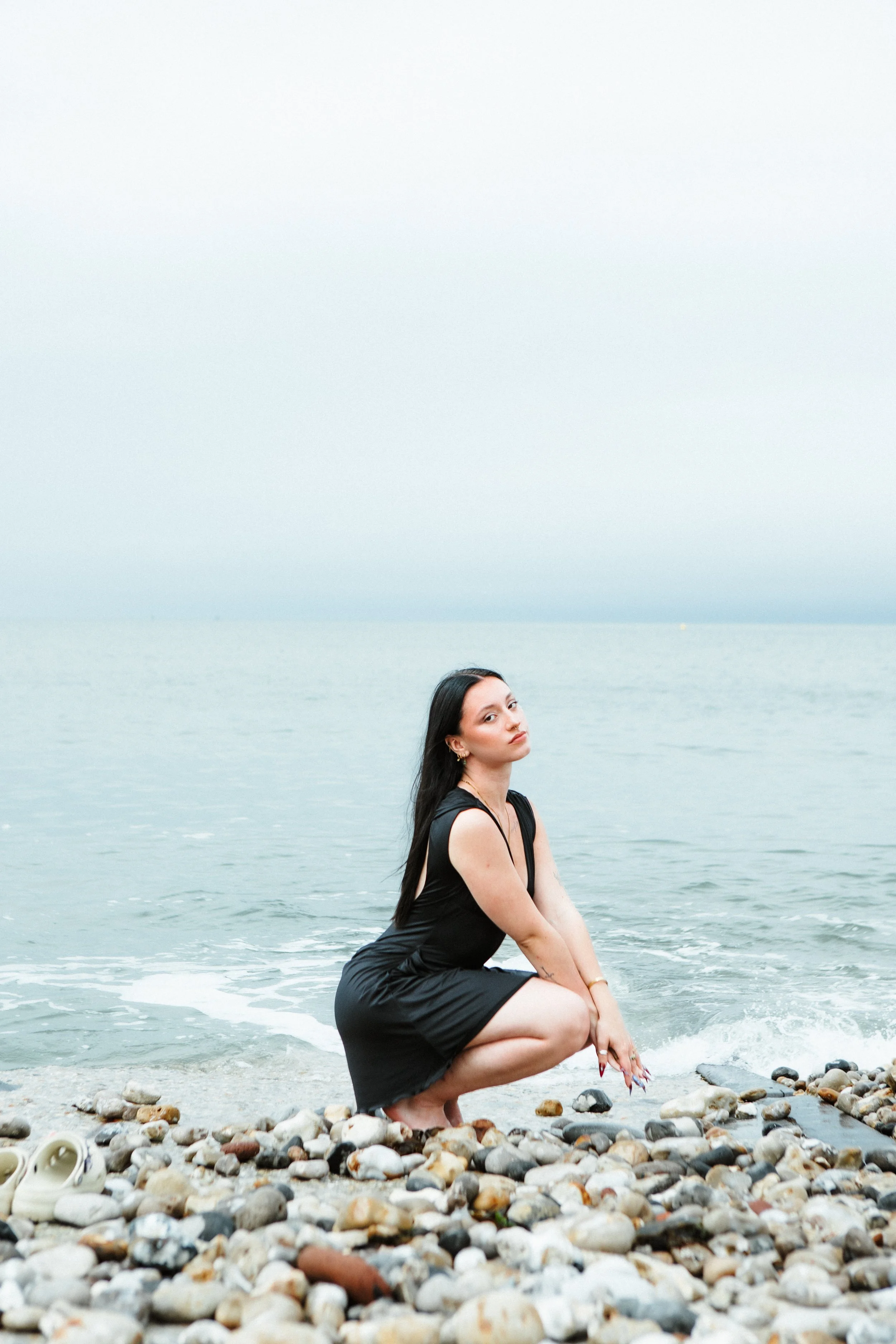 A woman with long black hair squatting on a pebble beach near the ocean, wearing a black sleeveless dress, with her arms resting on her knees, and a pair of white shoes placed on the rocks nearby, overcast sky in the background.