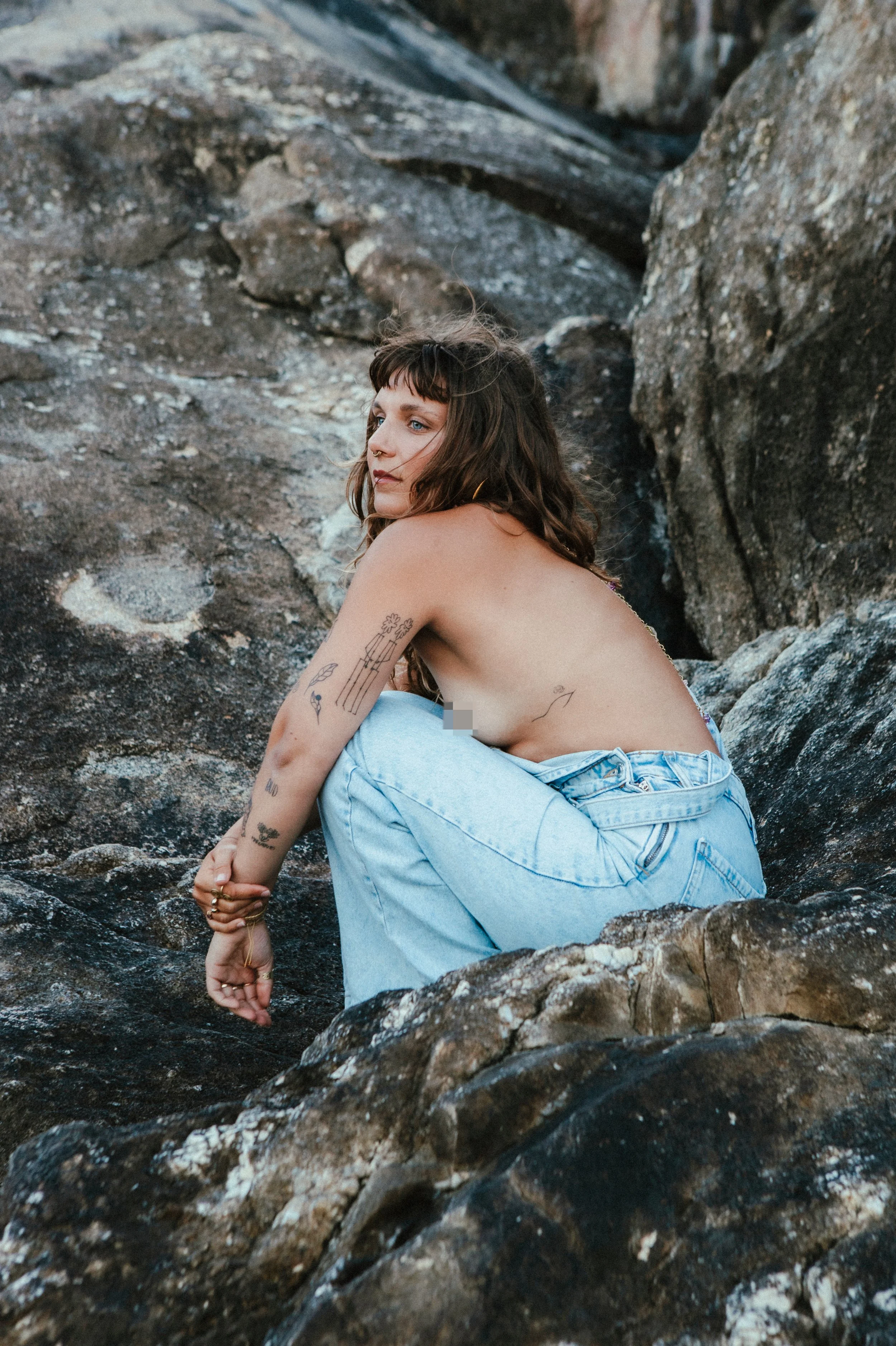 A young woman with tattoos and wavy brown hair sits among rocks near a small waterfall, looking contemplative.