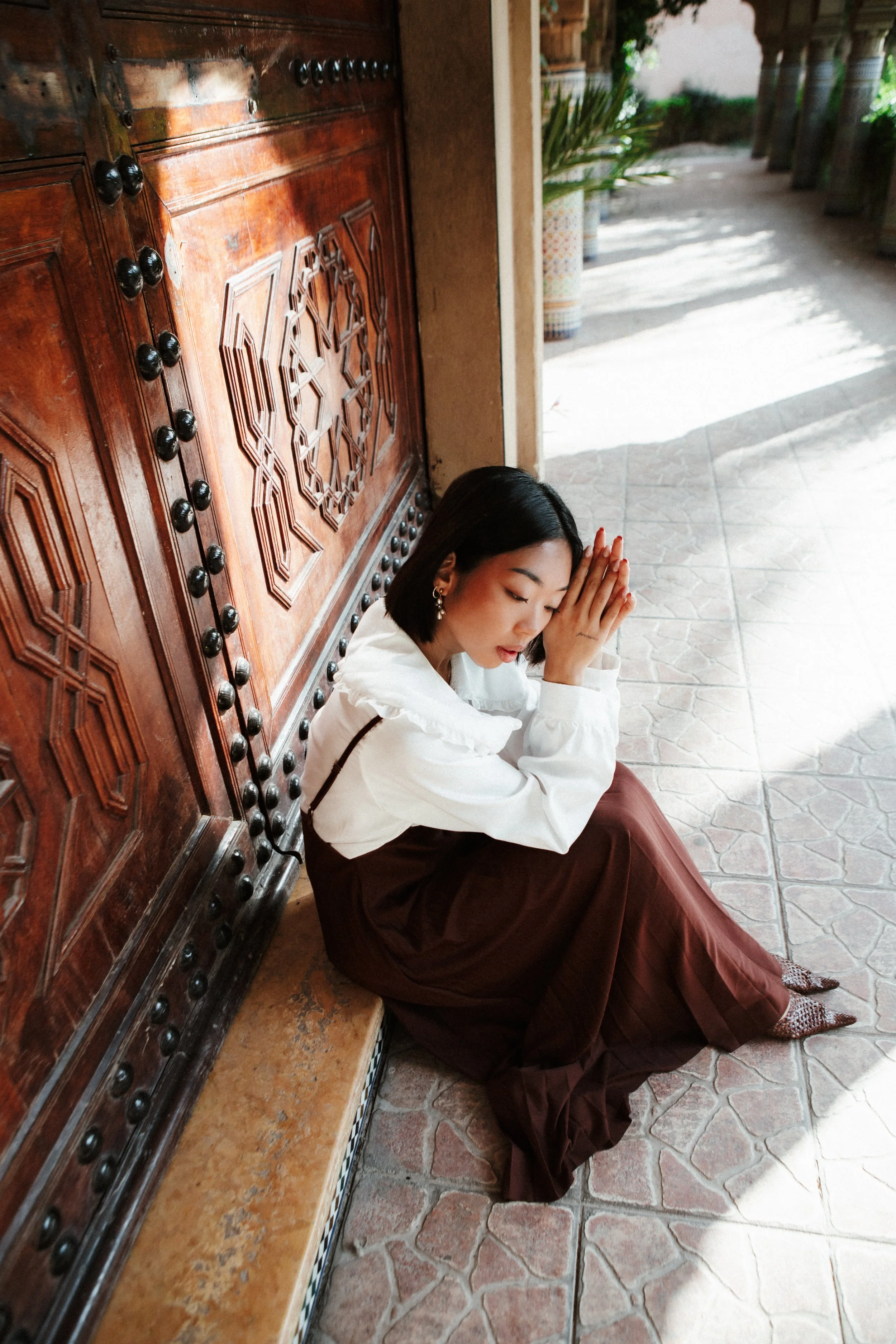 Young woman with short black hair and earrings, seated on a stone-tiled floor, praying with hands pressed together and head bowed, next to an ornate wooden door with carved designs and metal studs, in a sunny outdoor setting with potted plants in the