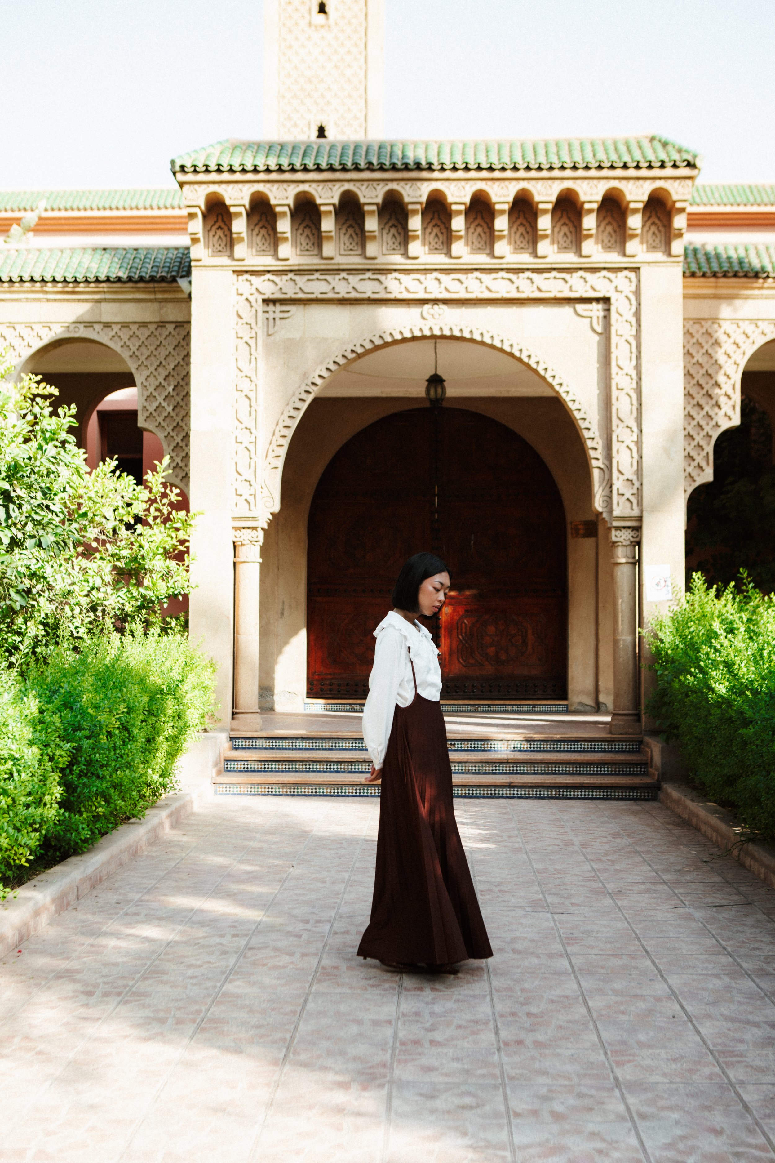 A young woman in white blouse and long brown skirt standing on tiled walkway in front of an ornate archway with green tiled roof and lush green bushes on sides.