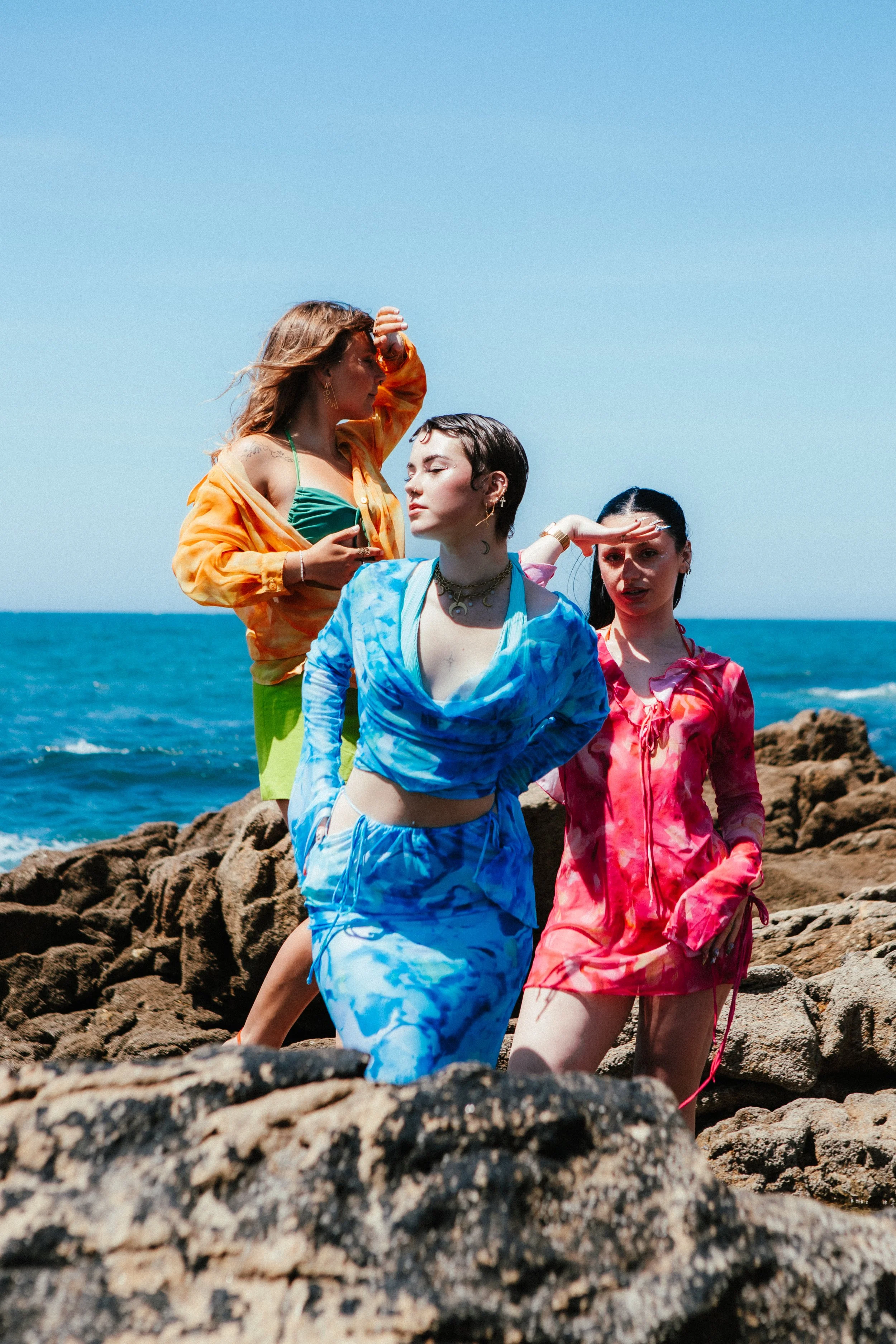 Three women in colorful, tie-dye beachwear stand on rocks by the ocean against a clear blue sky.