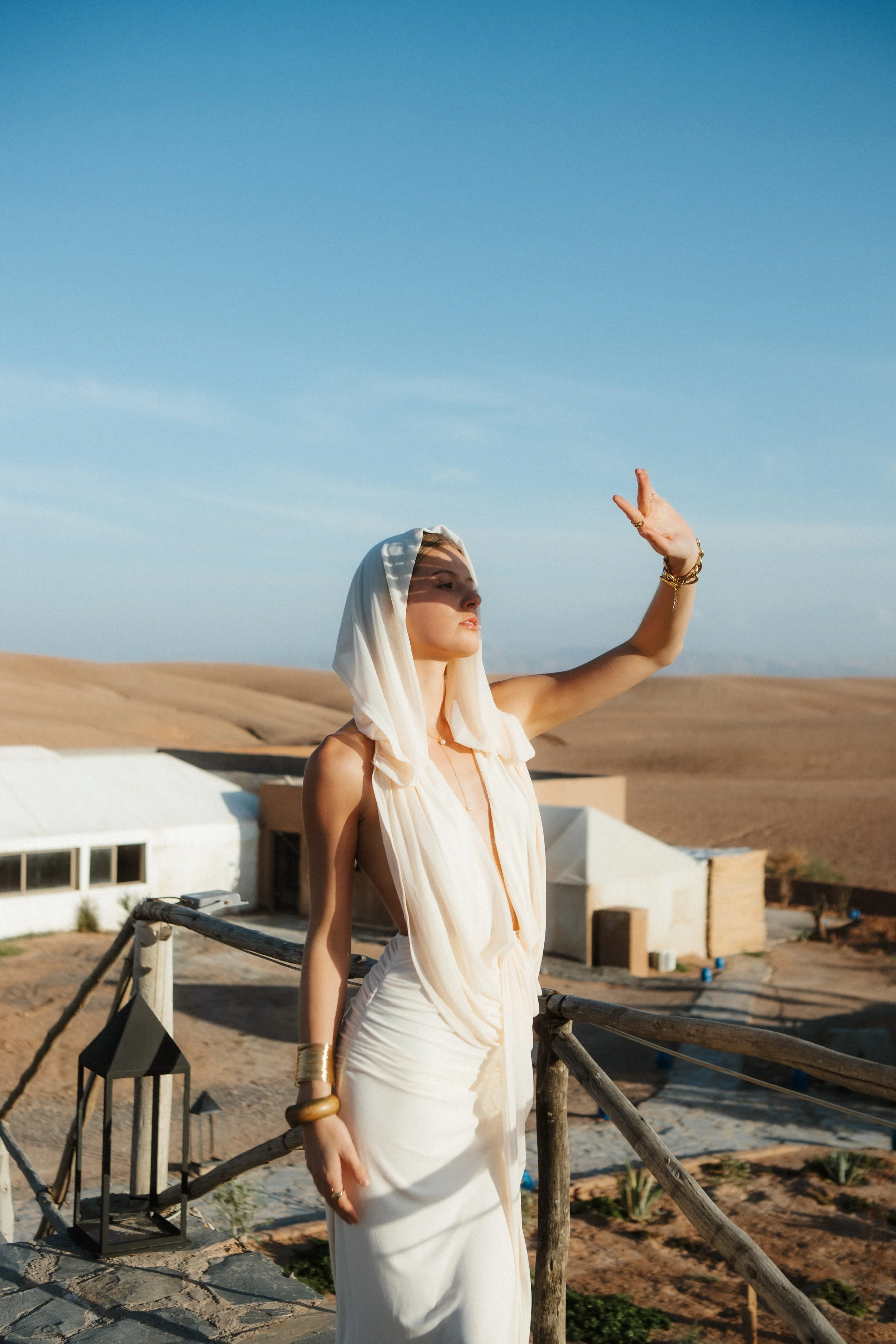A woman dressed in an elegant cream-colored dress with a hood, standing outdoors in a desert landscape with a clear blue sky, leaning on a rustic wooden fence and wearing gold jewelry.