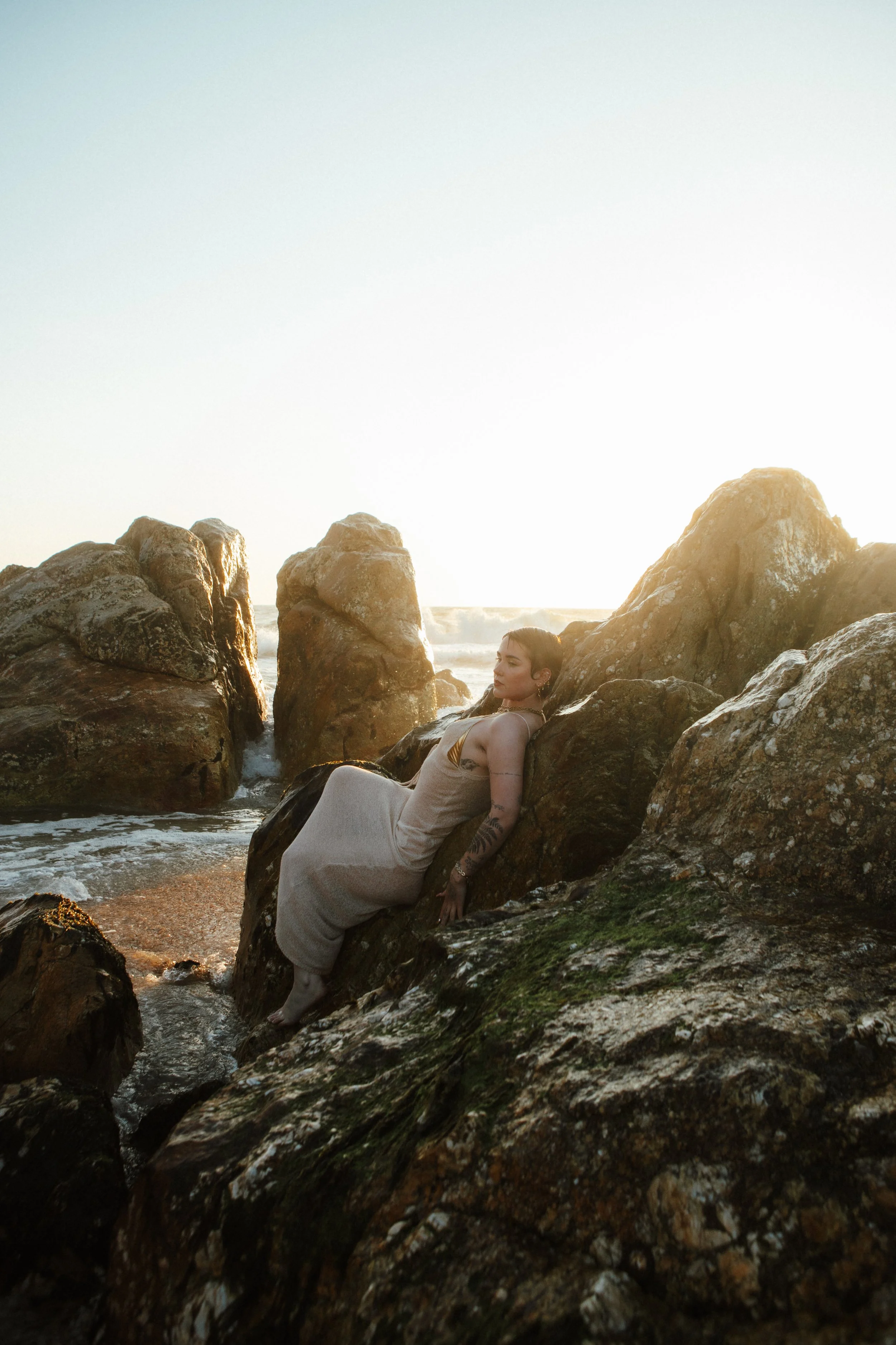 A woman in a light-colored dress relaxing on large rocks near the ocean during sunset, with waves crashing around her.