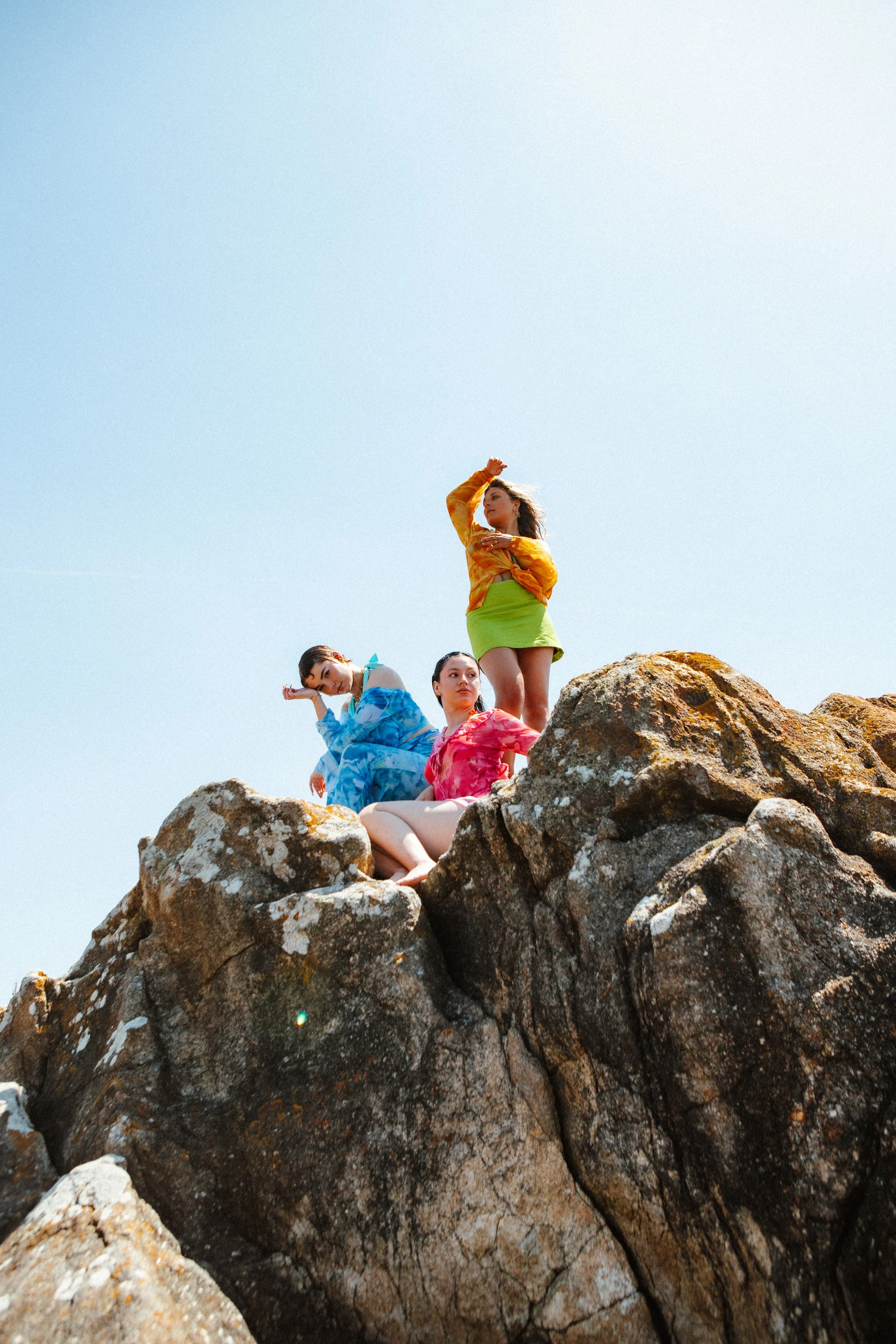 Three women in colorful outfits sitting and standing on large rocks outdoors against a clear blue sky.