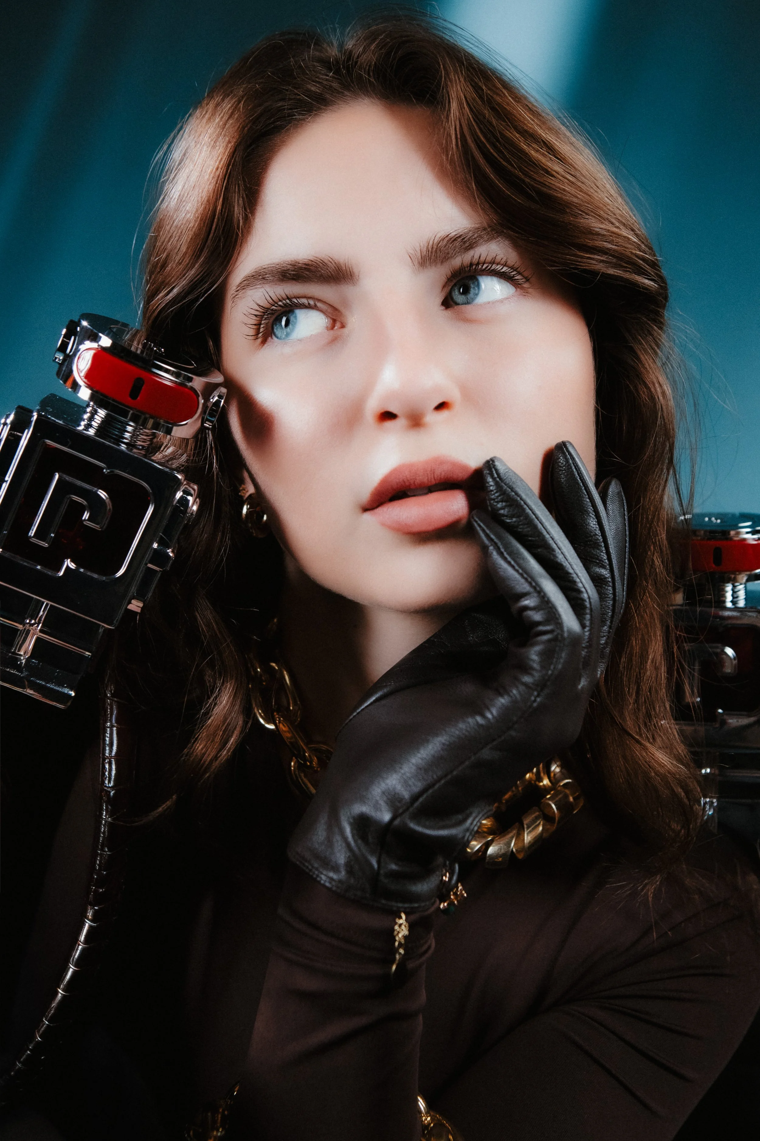 Close-up of a young woman with brown hair and blue eyes, wearing black leather gloves, gold jewelry, and holding a camera with a red and black strap, against a blue background.
