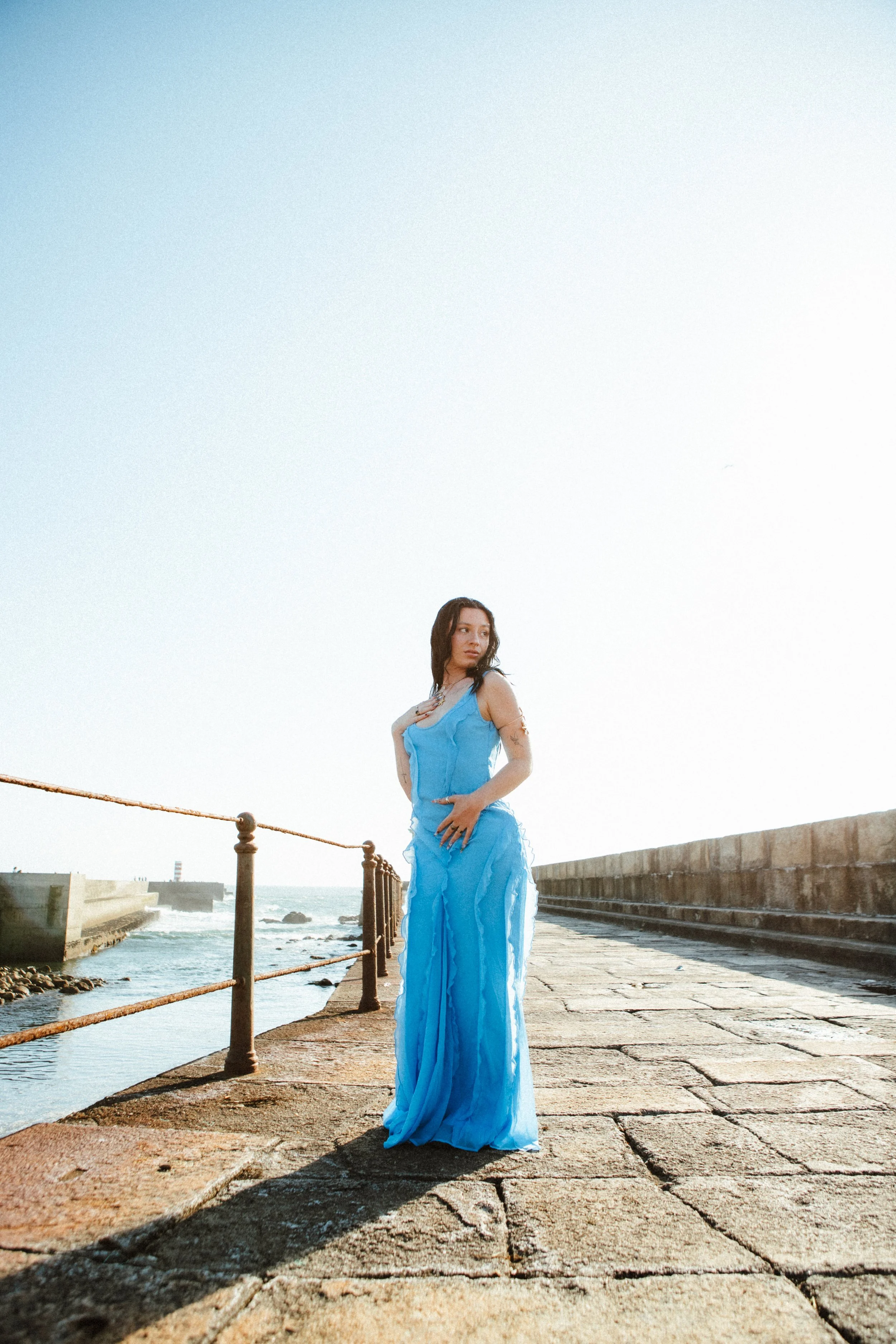 Woman in a flowing blue dress standing on a stone pier by the ocean during sunset, with a lighthouse in the distance.