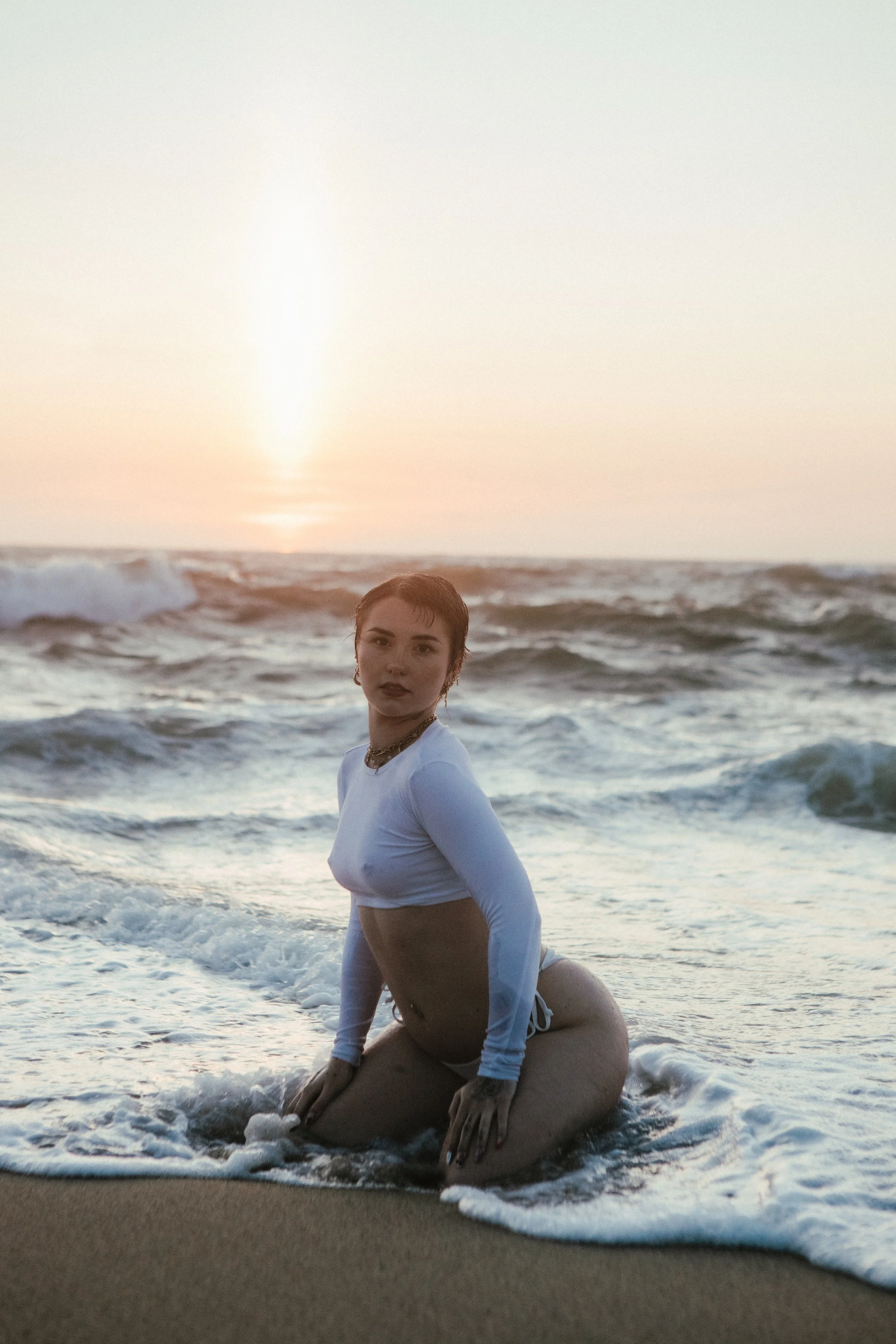 A woman with short dark hair and jewelry is kneeling on the beach in the ocean at sunset, with waves around her and the sun low in the sky.