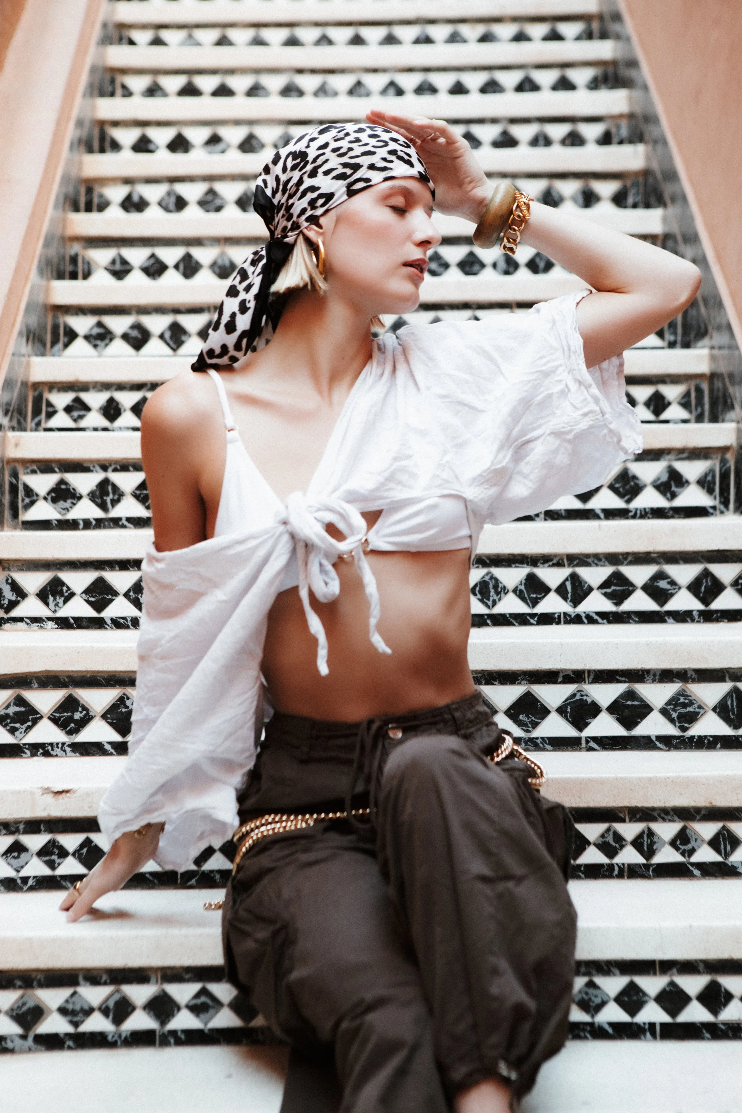A woman with blonde hair, wearing a leopard print headscarf, gold hoop earrings, multiple bracelets, a white tied crop top, and dark pants, sitting on patterned stairs with her eyes closed and hand on her forehead.