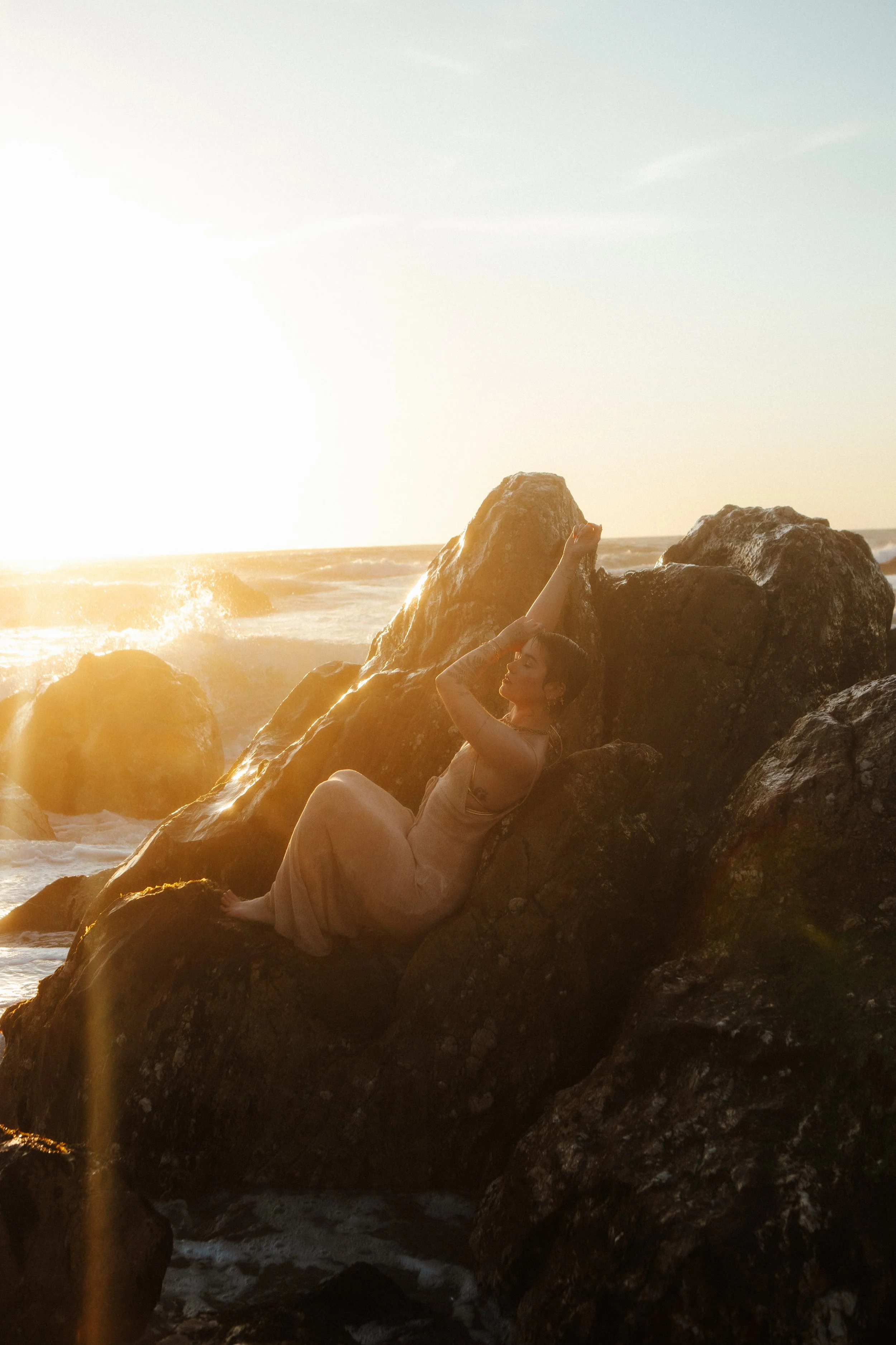 A woman in a long beige dress sitting on large rocks at a beach during sunset, with waves crashing behind her and the sun low in the sky.