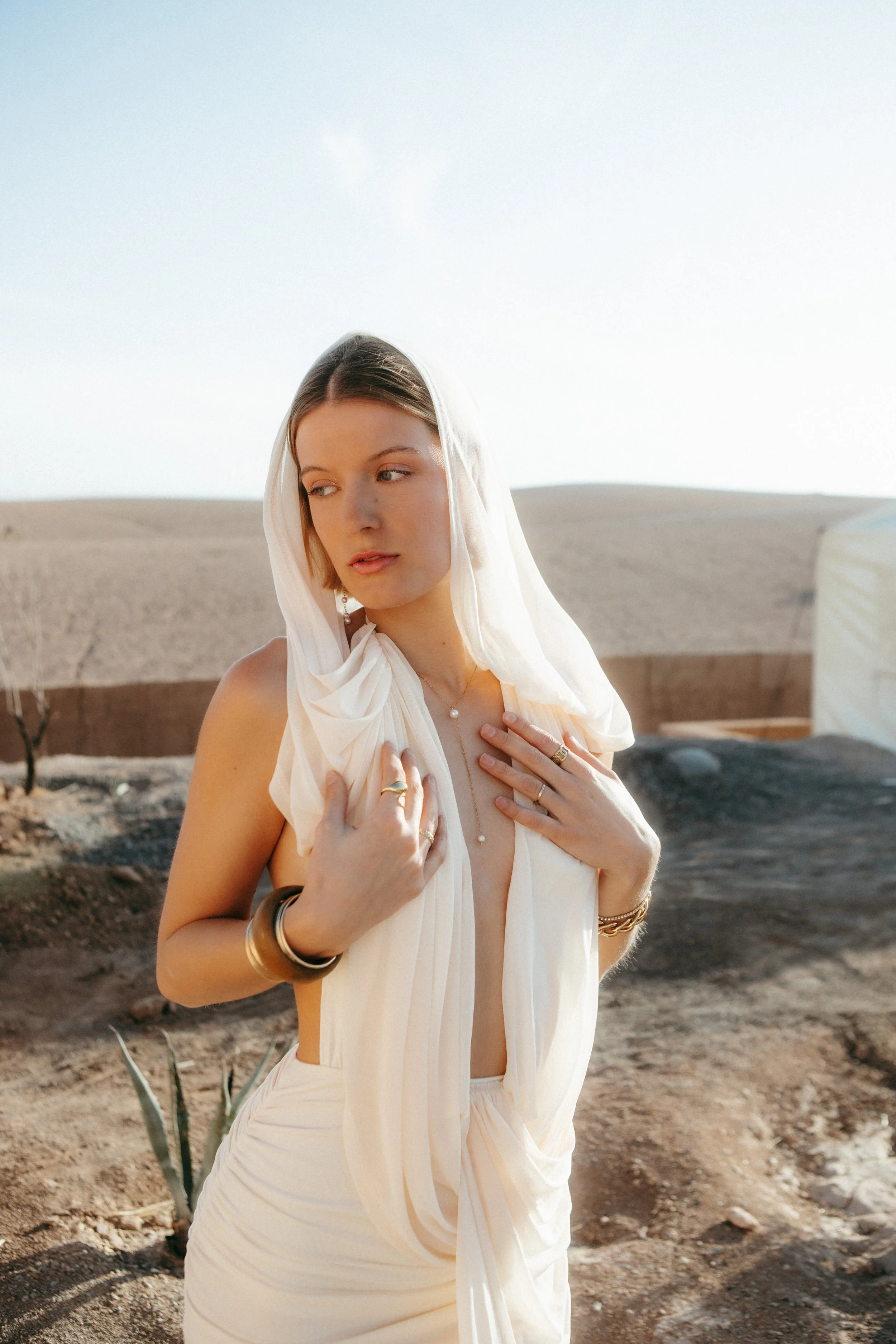 A woman with short hair and fair skin wearing a white headscarf and flowing cream-colored dress in a desert landscape with cacti and a tent in the background.
