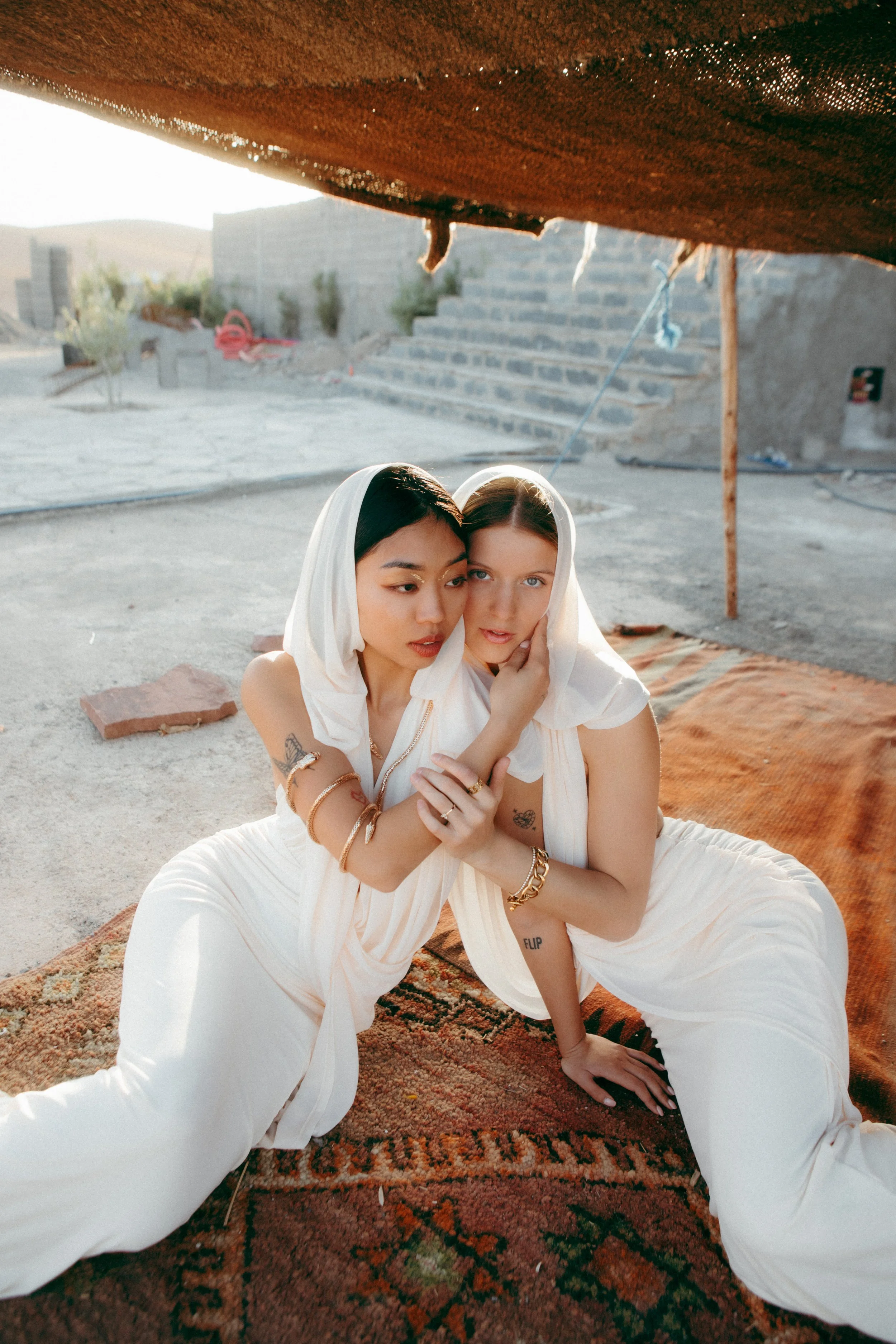 Two women sitting on a patterned rug underneath a makeshift canopy in a desert setting with stairs and sand in the background. One has dark hair and the other has light hair, both are in white clothing and are embracing each other.