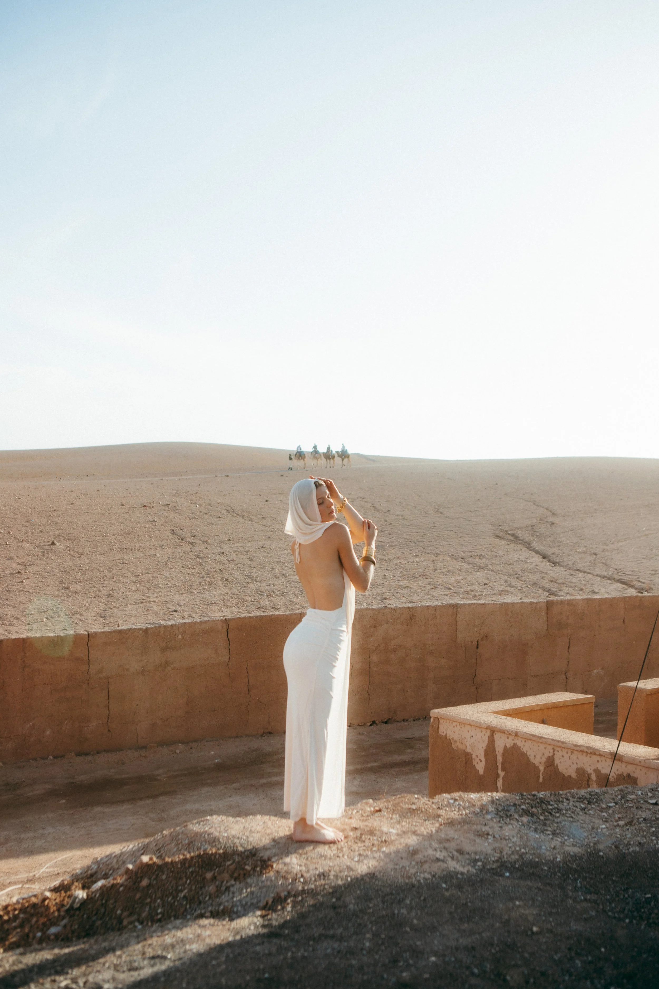 A woman with a white headscarf and white high-waisted pants standing outdoors in a desert-like area during daytime. In the background, there is a sandy landscape and a caravan of camels with riders.
