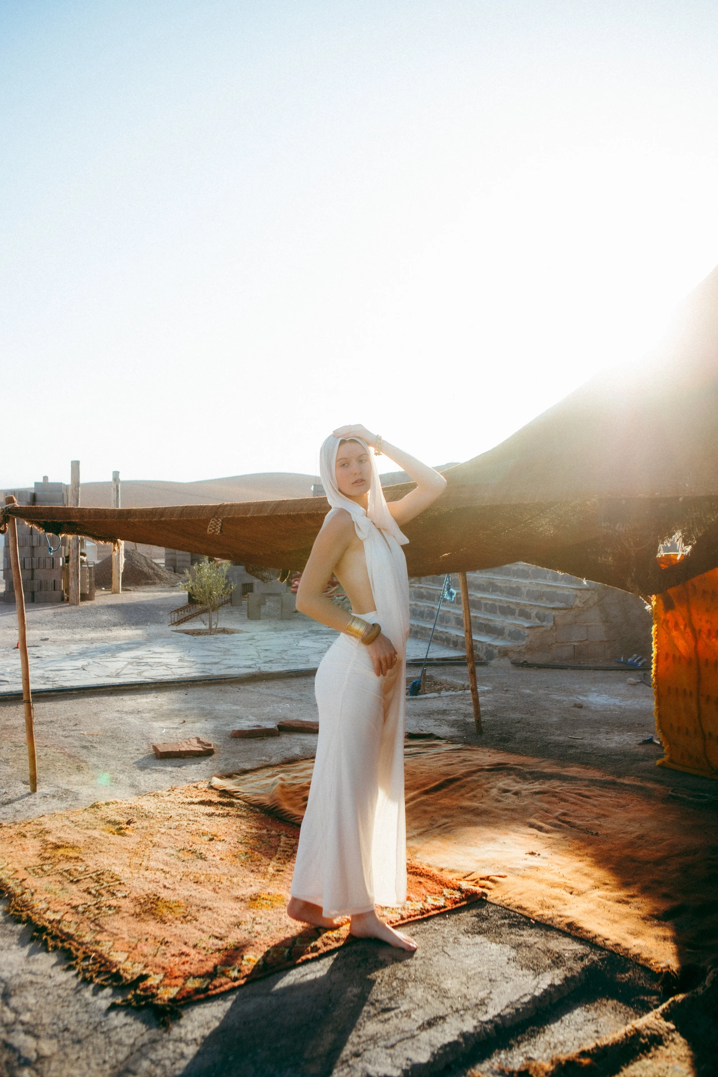 A woman in a cream-colored dress standing barefoot on an ornate carpet outdoors at sunset. She is holding her head with one hand, looking directly at the camera, with a beige headscarf draped over her shoulders. There are blankets or rugs on the grou