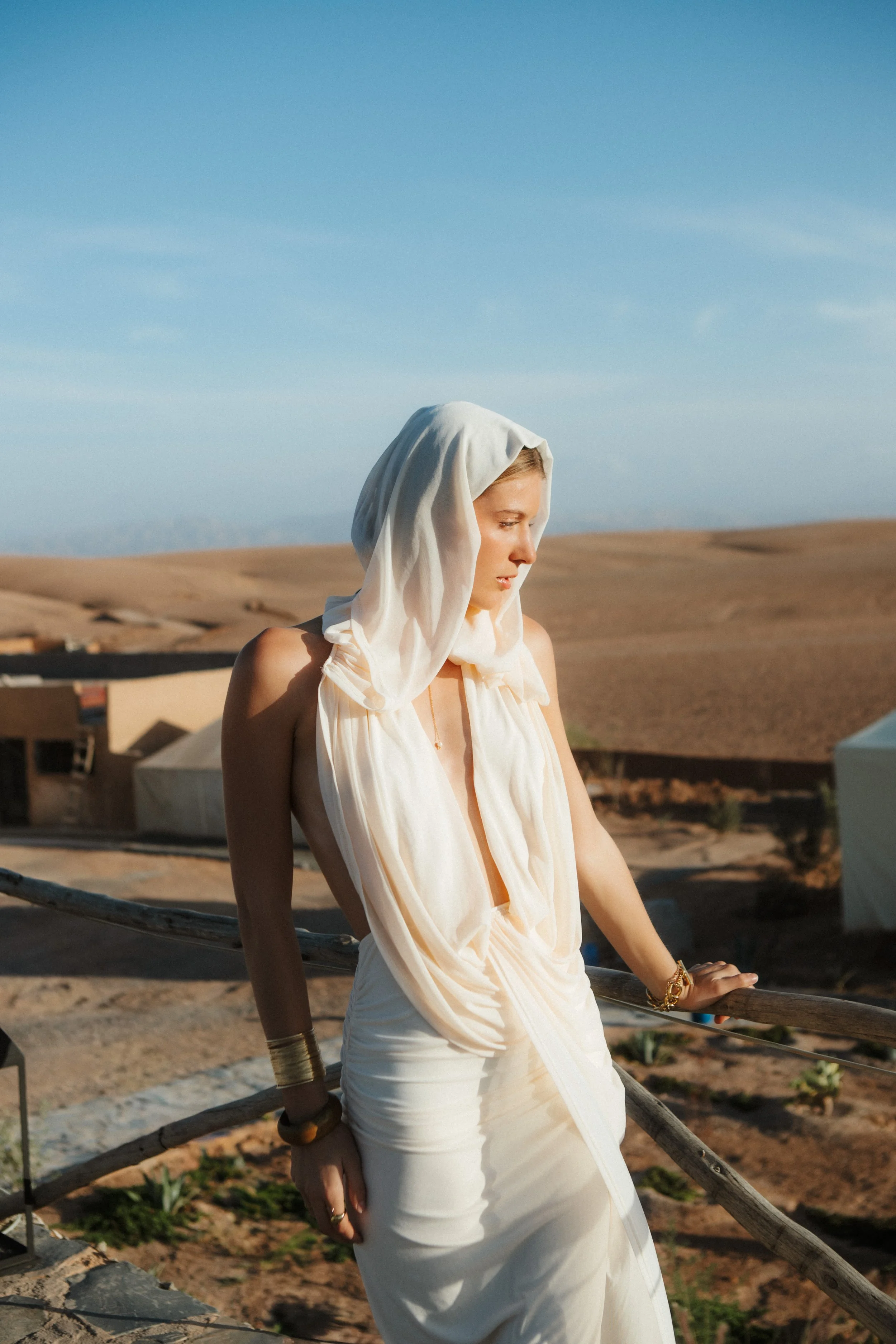 A woman wearing a white dress and a head covering, standing outdoors in a desert landscape with hills under a blue sky.
