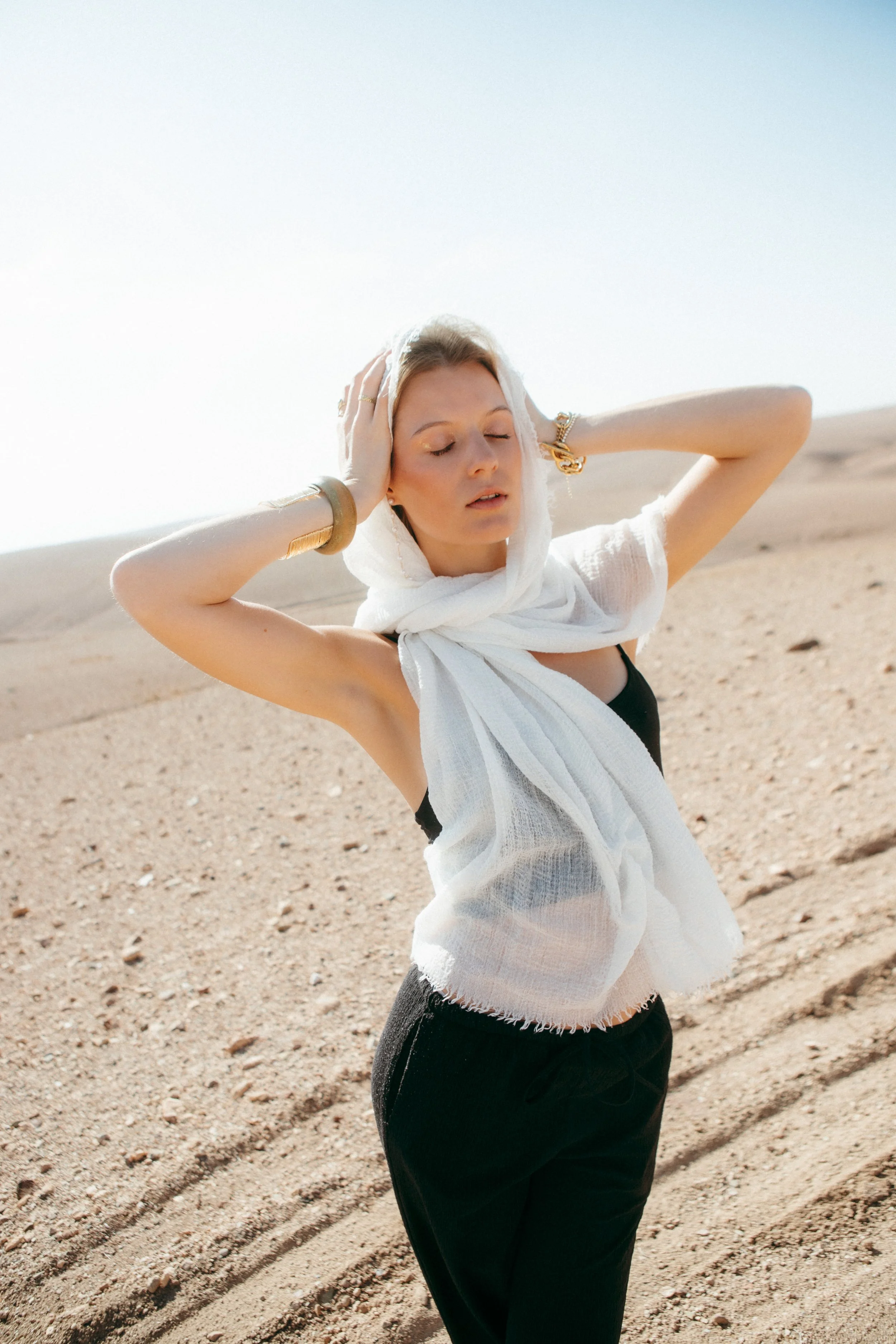 A woman in a black top and black pants with a white headscarf and large gold jewelry stands in a sandy outdoor setting with her eyes closed and hands in her hair.