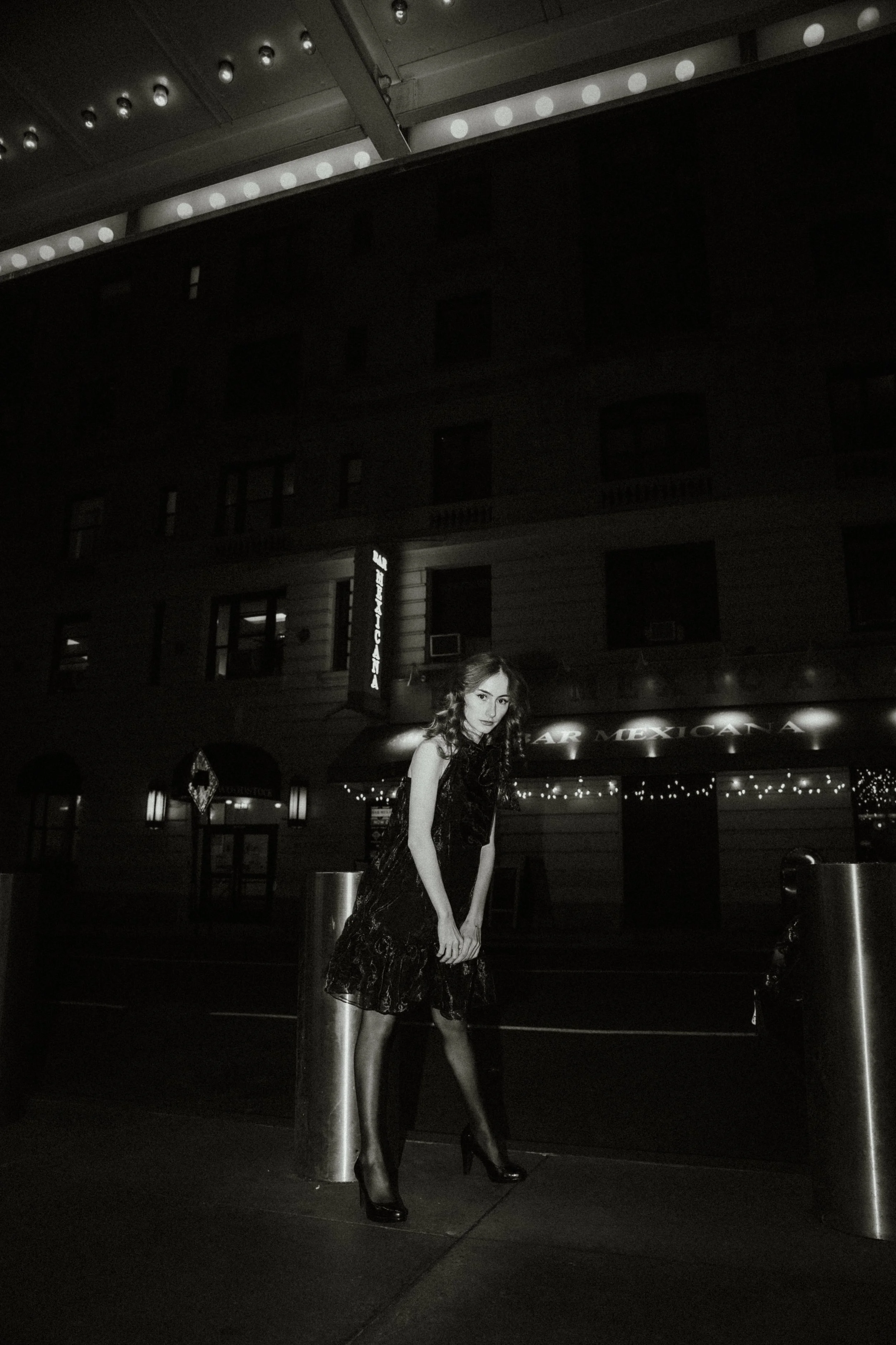 A woman wearing a black dress and high heels standing outside at night near a metal post, with a building with lit windows and a sign that reads 'Bar Mexicana' in the background.