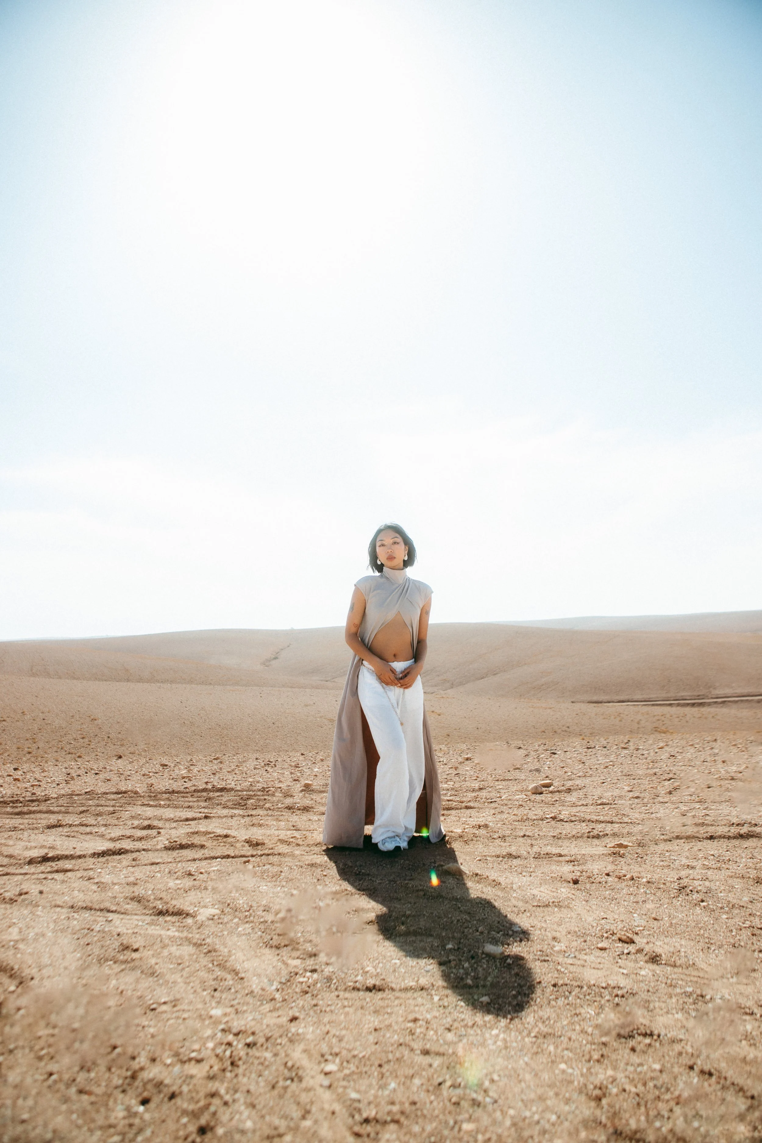 A woman standing in a desert landscape with sand dunes and a clear sky, wearing a sleeveless top and long skirt, looking into the distance.