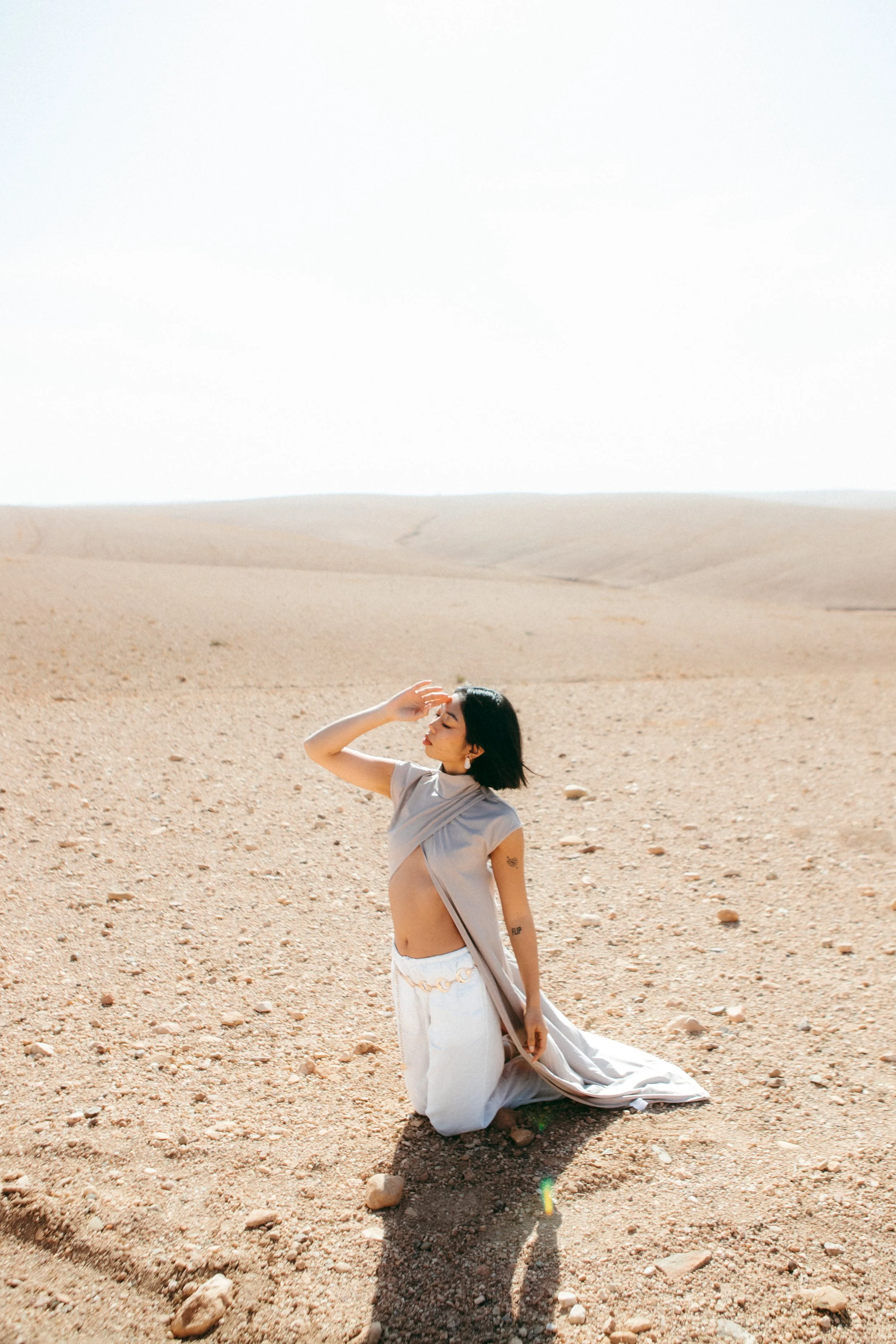 A woman kneeling on desert sand, wearing a beige top and white skirt, with her hand raised to her forehead, in a vast, arid landscape under bright sunlight.
