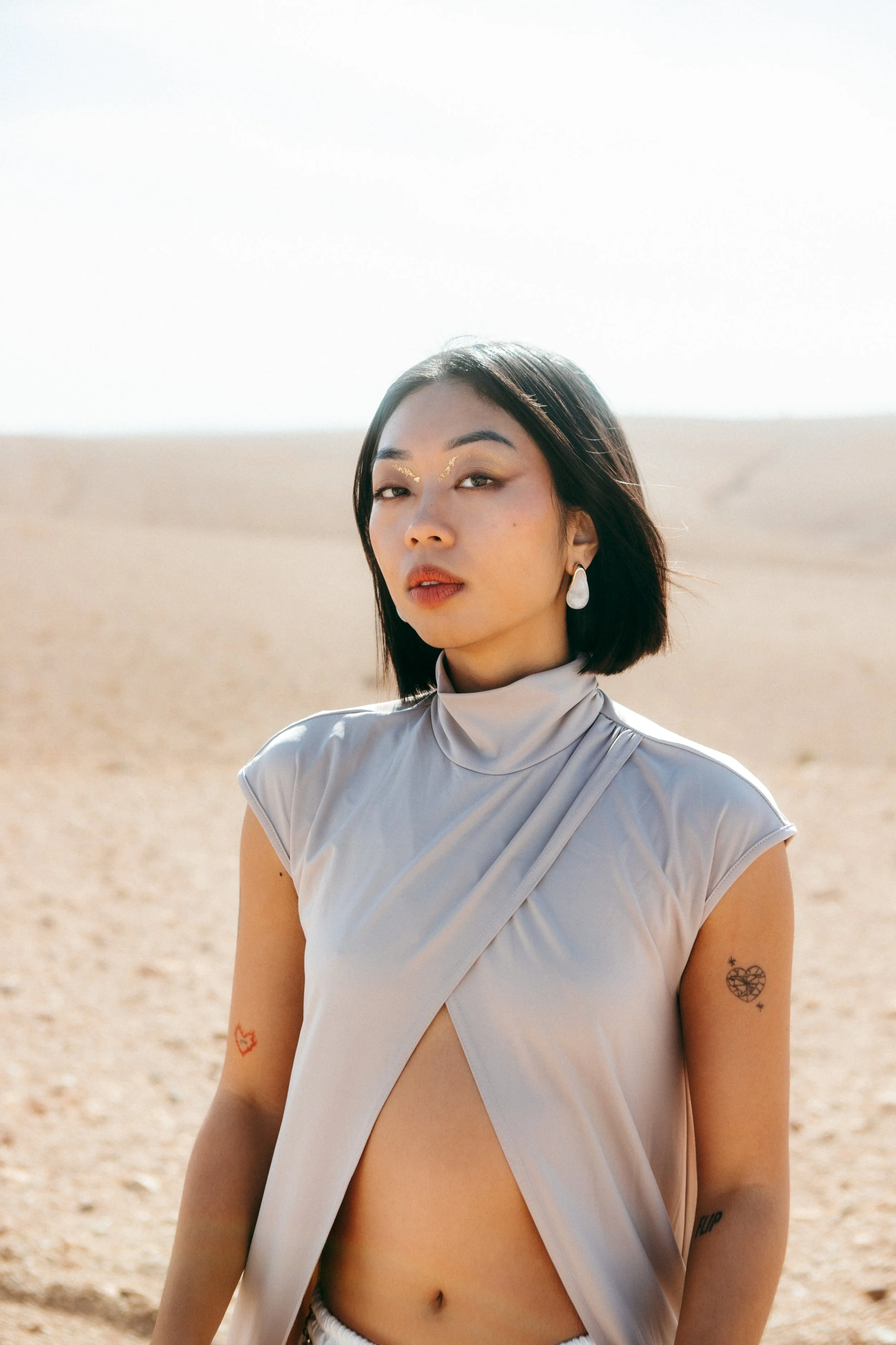 A woman with short black hair and earrings stands in a desert landscape with a bright sky in the background.