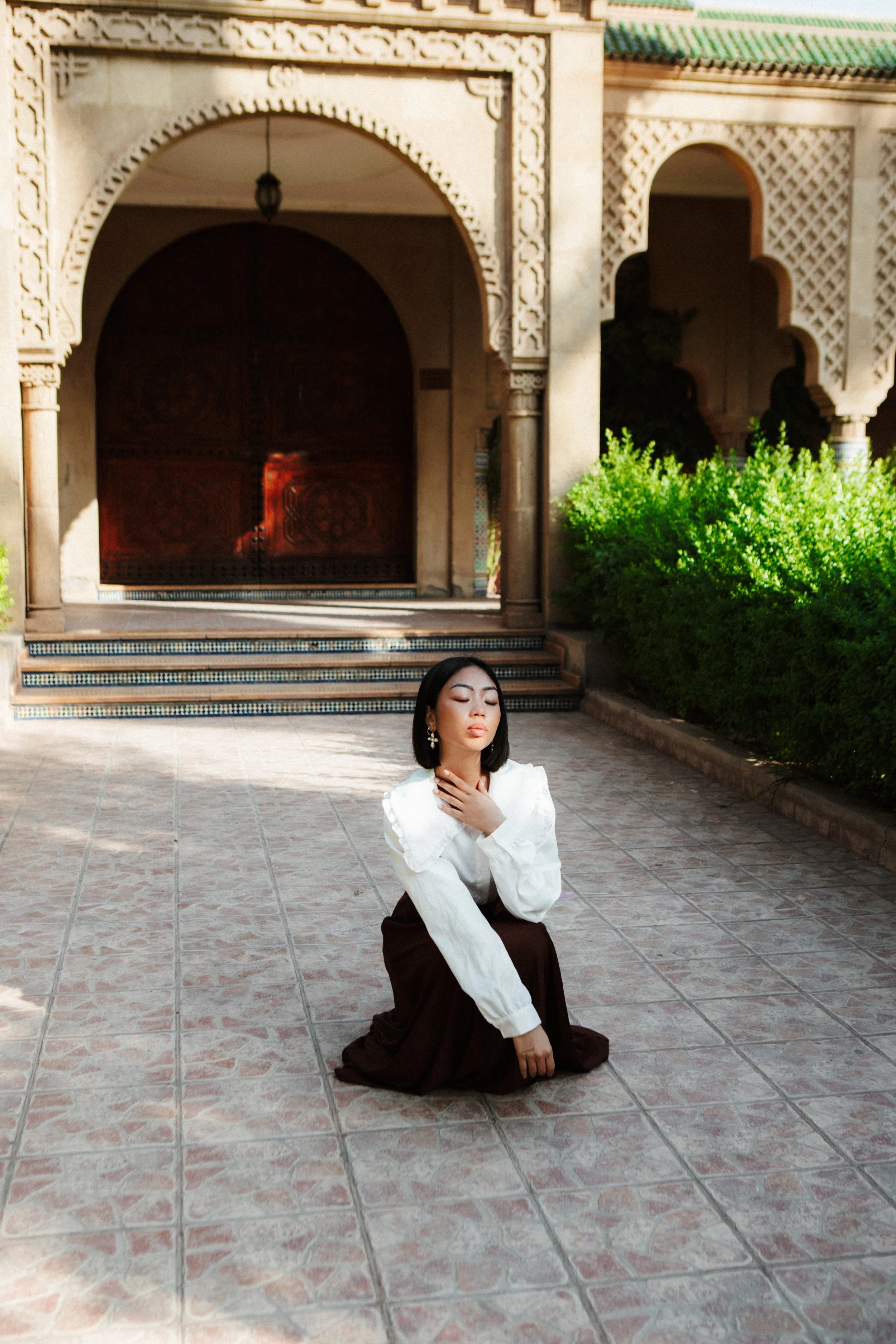 A woman in a white blouse and dark skirt is kneeling on a tiled patio with her hands gently touching her neck, eyes closed, and surrounded by lush green shrubbery, with traditional ornate architecture in the background.