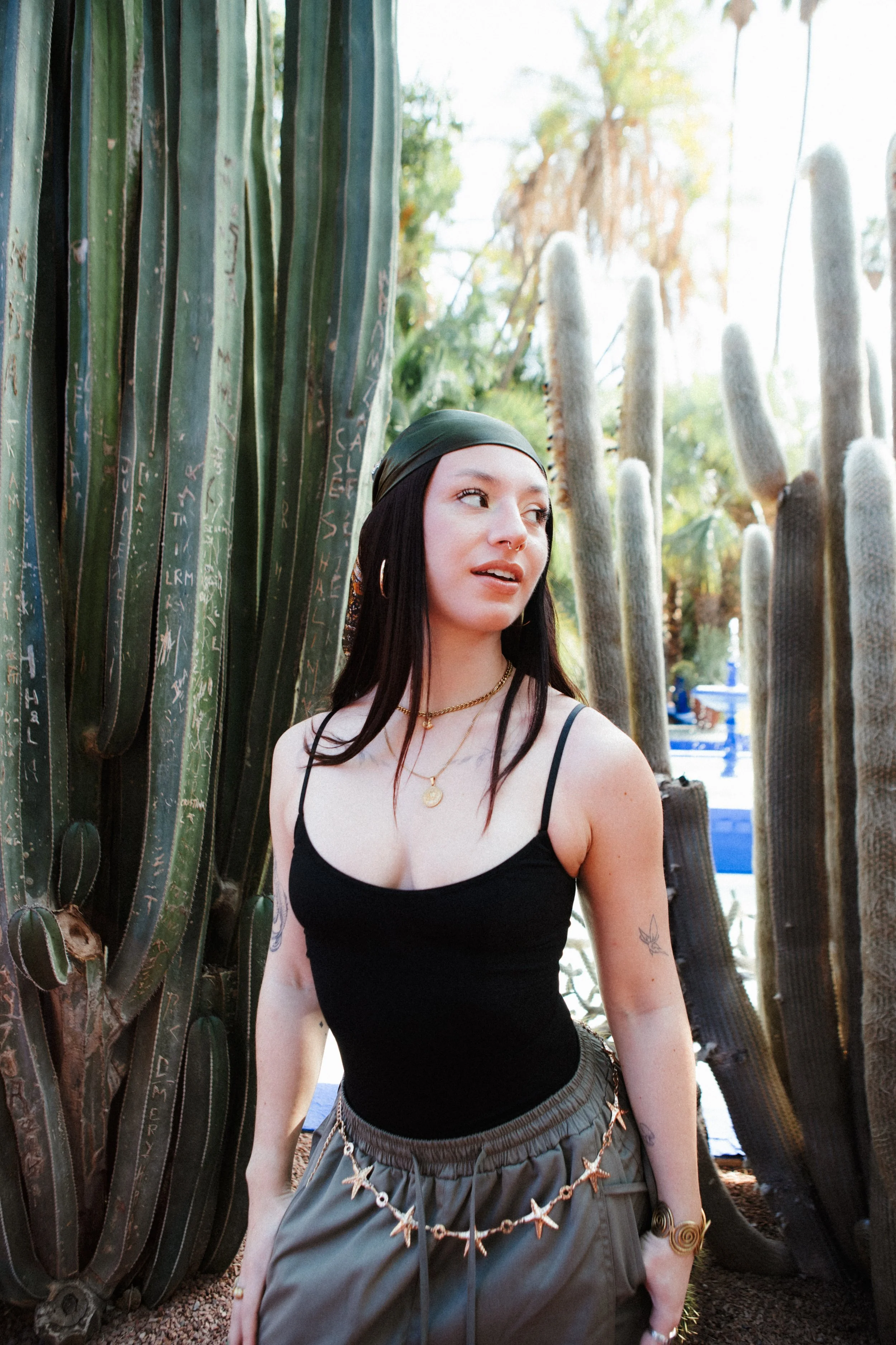 A young woman with dark hair and tattoos on her arms standing among tall cacti. She is wearing a black spaghetti strap top, olive green pants with a starfish chain belt, gold jewelry, and a dark headscarf, looking to the side.